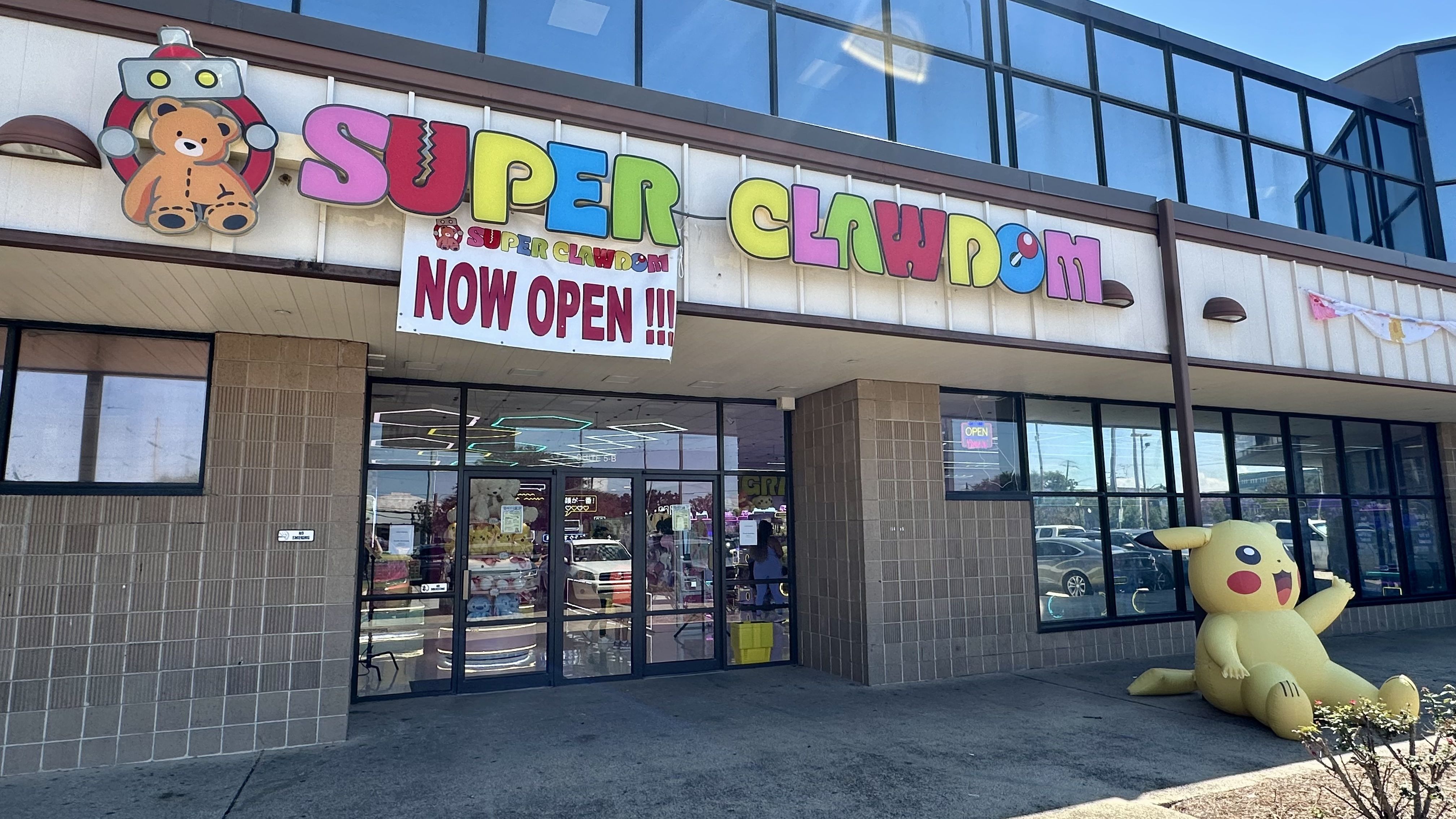 Exterior of Super Clawdom arcade with colorful sign, teddy bear logo, "NOW OPEN!!!" banner, glass entrance, and large inflatable Pikachu figure outside under blue sky.