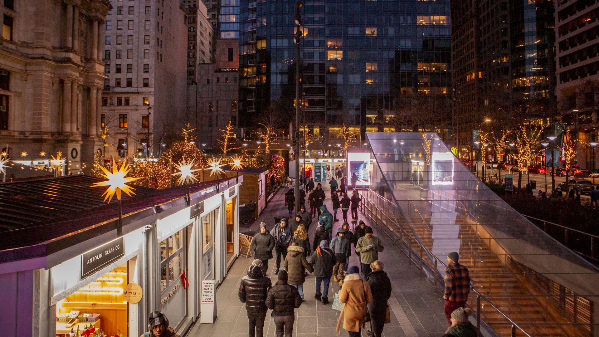 Evening city scene with people walking near lit holiday market stalls and a glass subway entrance, surrounded by tall buildings and tree lights.