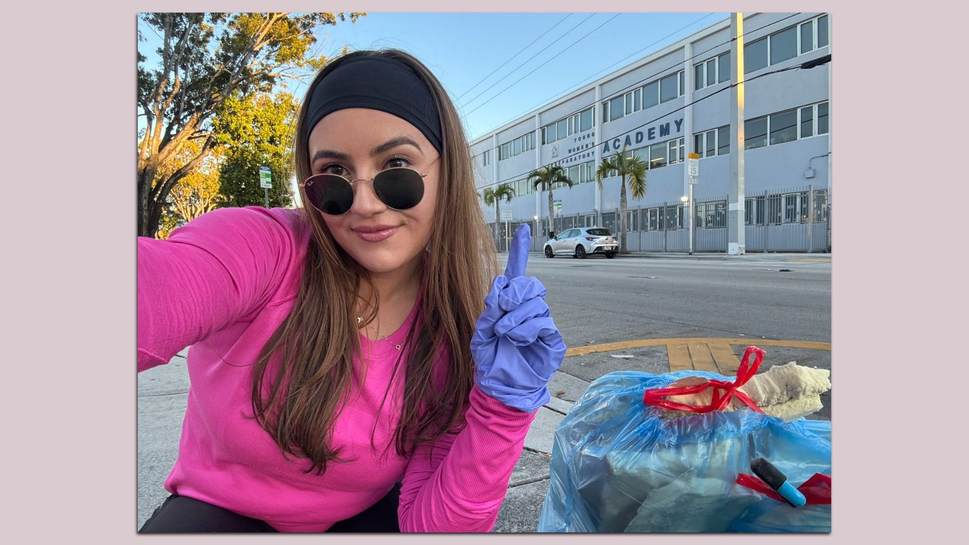 Young woman wearing a black headband, sunglasses, pink top, and purple gloves takes a selfie on a street; a blue trash bag is in the foreground and a gray building labeled "ACADEMY" in the background.