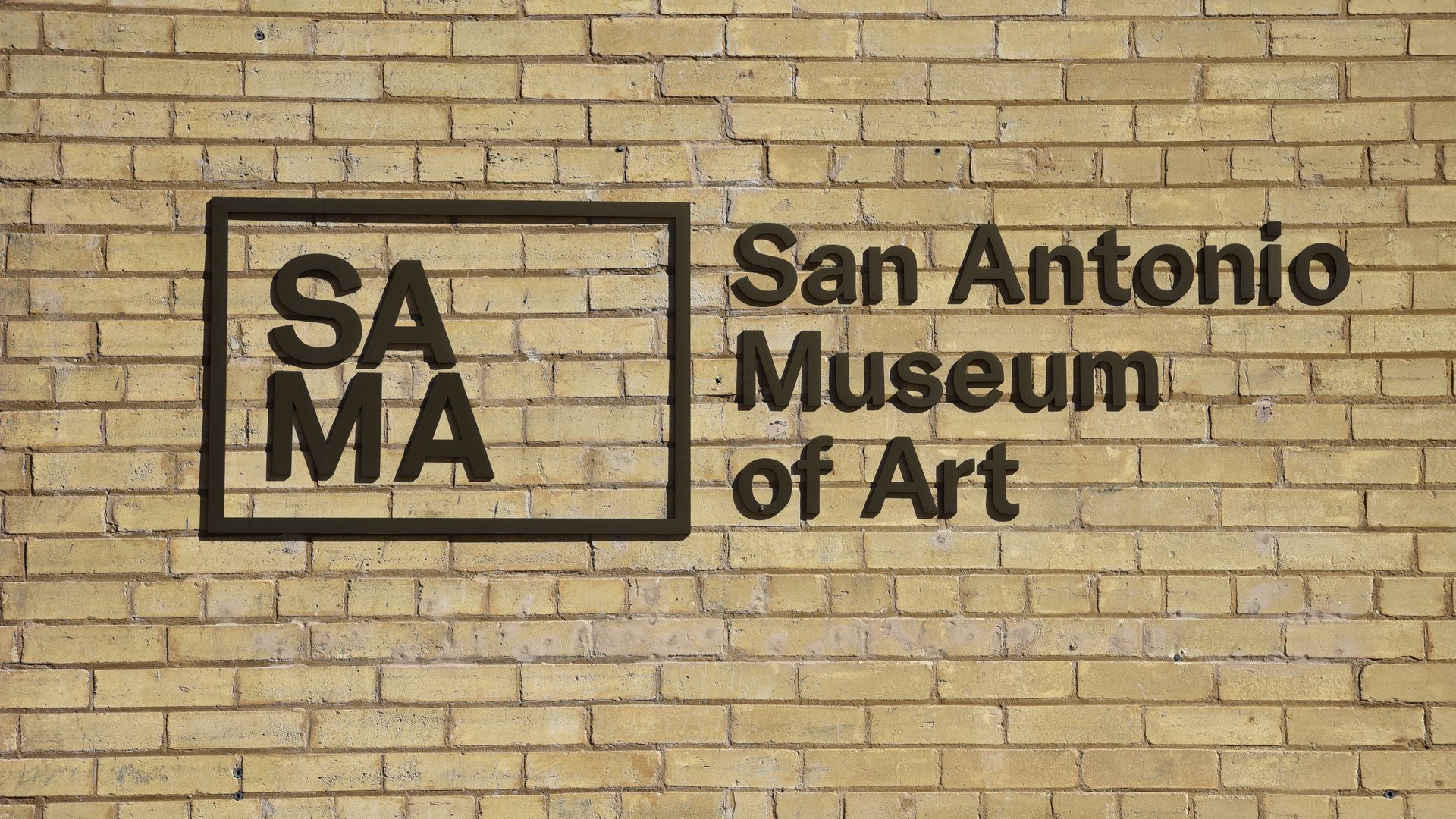 A logo that reads "SAMA" inside a box next to text that reads "San Antonio Museum of Art" against a light-colored brick wall on the outside of the museum.