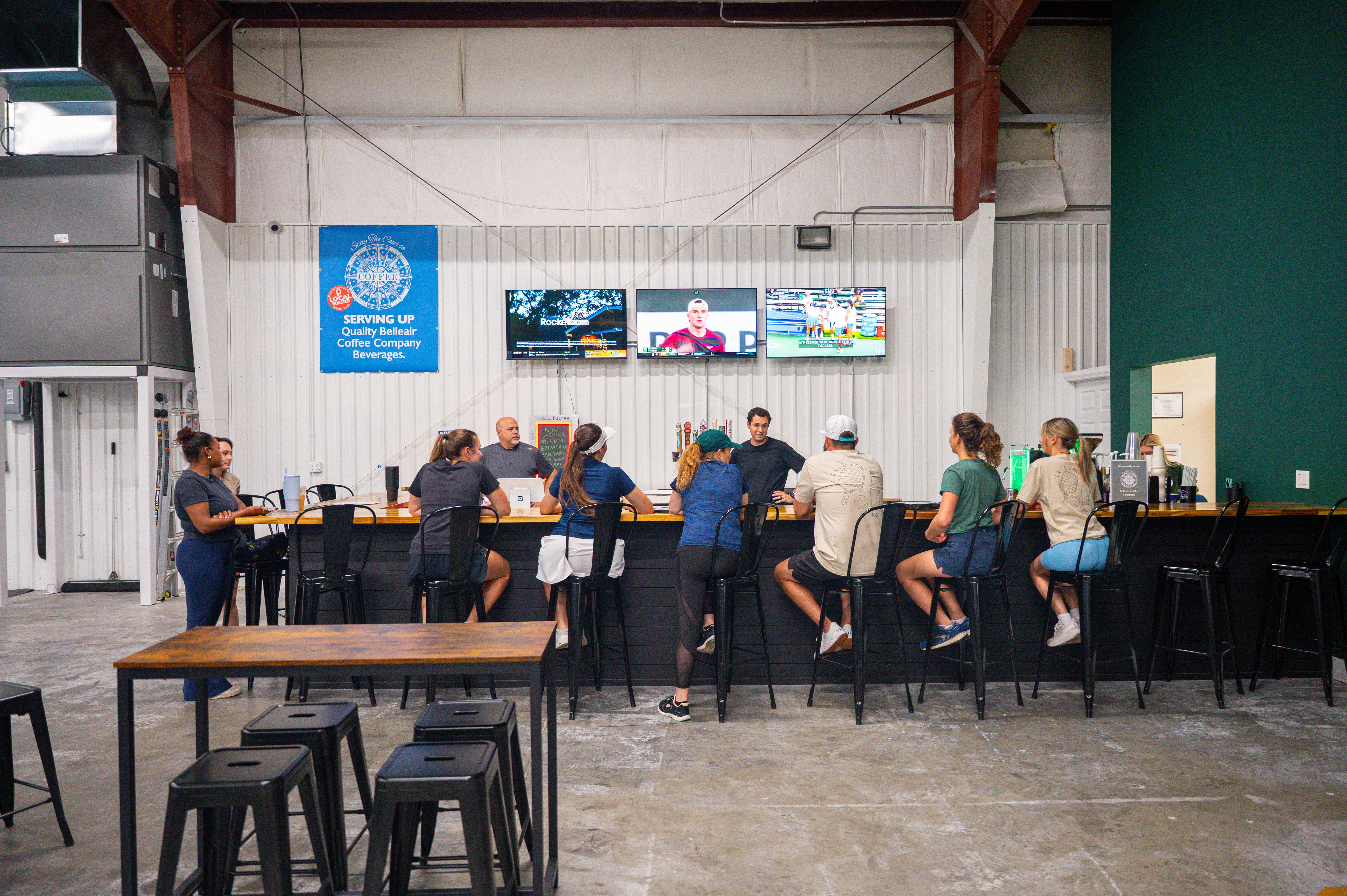 Group of people sitting and standing around a wooden bar counter in an industrial-style cafe with three TVs on the white wall showing sports, and a blue sign advertising coffee.