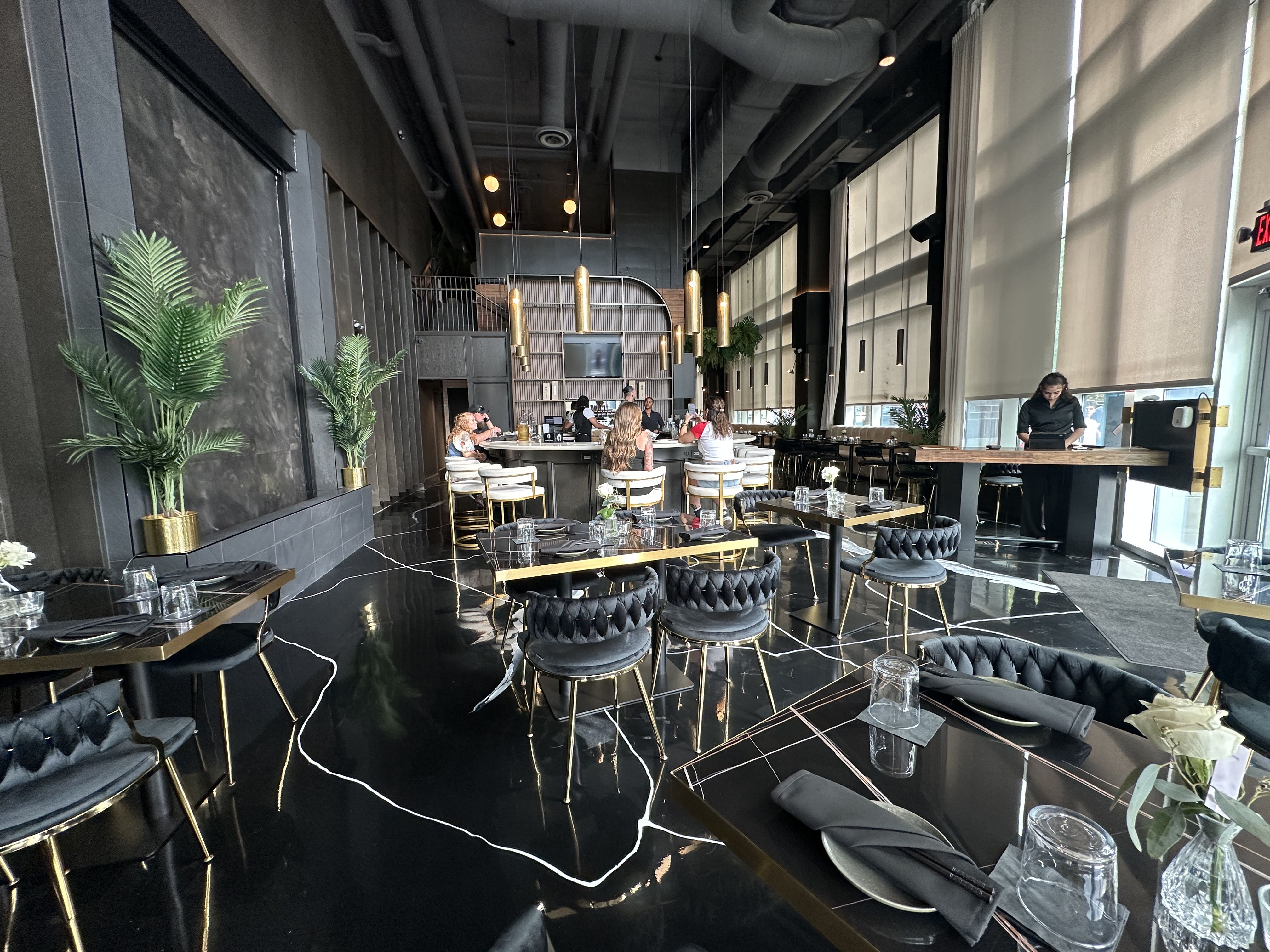 LILA. Upscale restaurant interior: black marble floor, gold accents, pendant lights, dark walls, large windows, plants; diners at round tables and a staff member at a right-side counter.