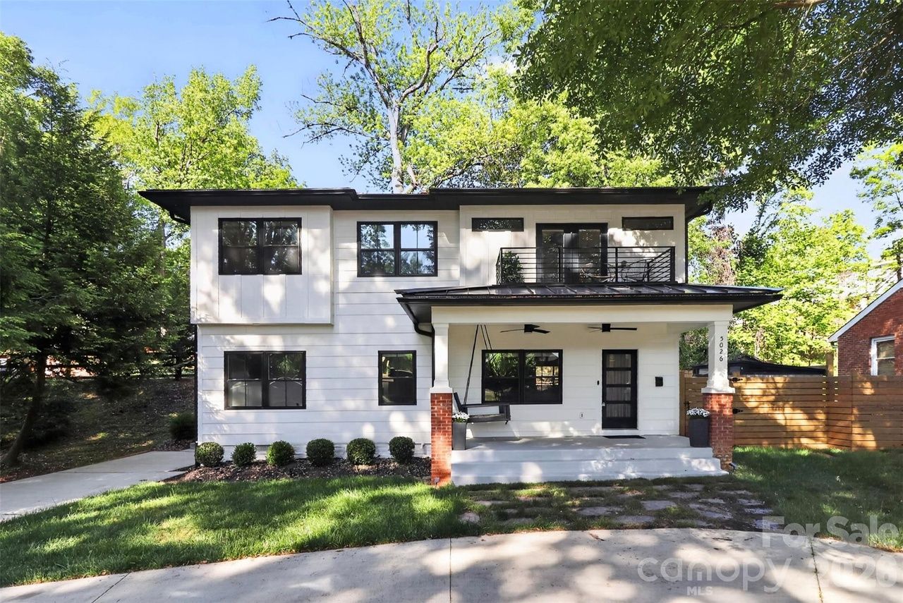 A two-story white modern home with black trim, a covered front porch supported by brick columns, black-framed windows, a small second-floor balcony, and a green lawn with surrounding trees; "For Sale" watermark.