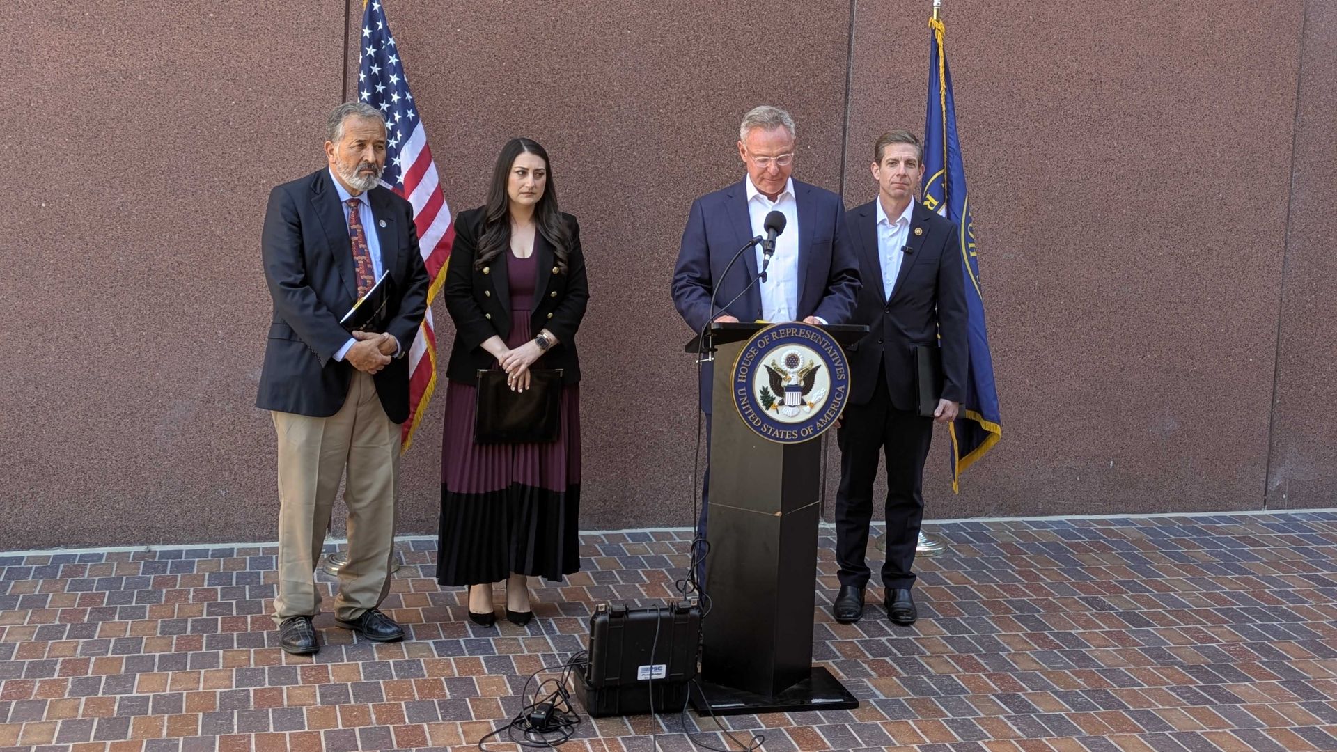Four people stand at a podium with the House of Representatives seal, flanked by US and state flags, against a brown wall and brick ground during a formal press event.