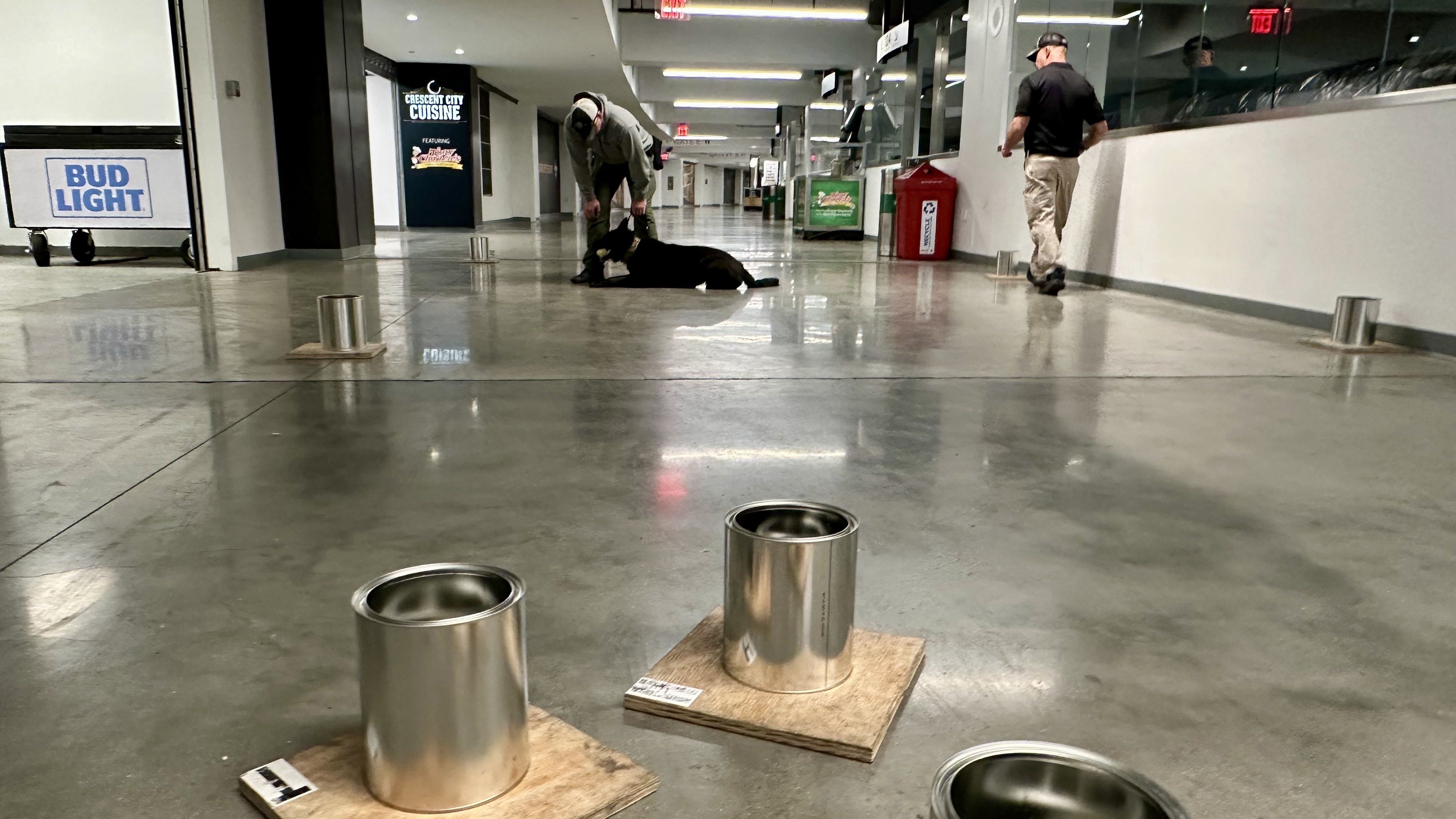 Indoor corridor: a man in a gray hoodie kneels beside a black dog on a polished concrete floor; another man walks away, while metal cans on boards line the hallway; a Bud Light sign is left.