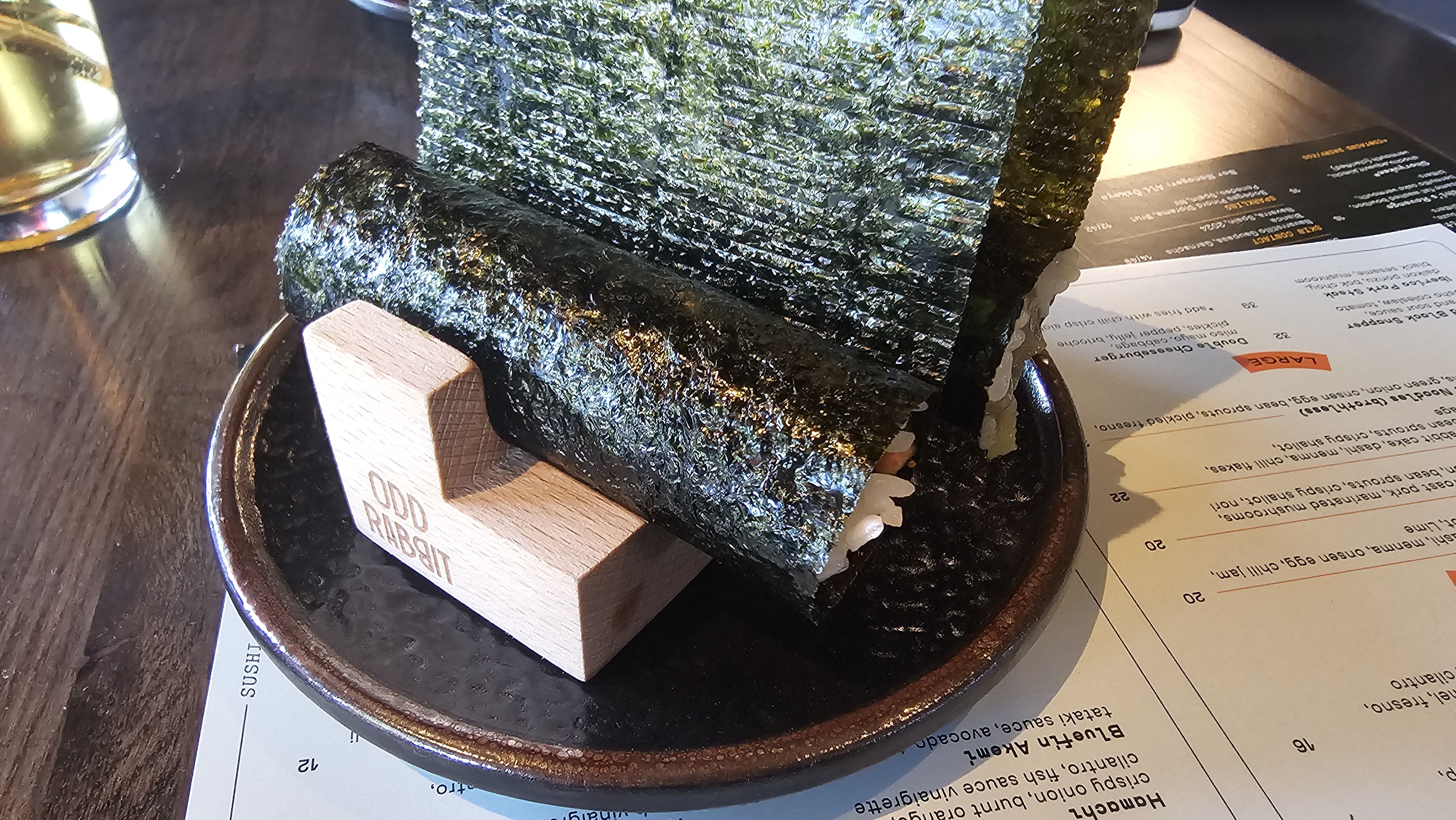 Round plate with dark, glossy nori sheets stacked upright; a wooden block rests at the base. A drink and a menu are visible on a wooden table in the background.