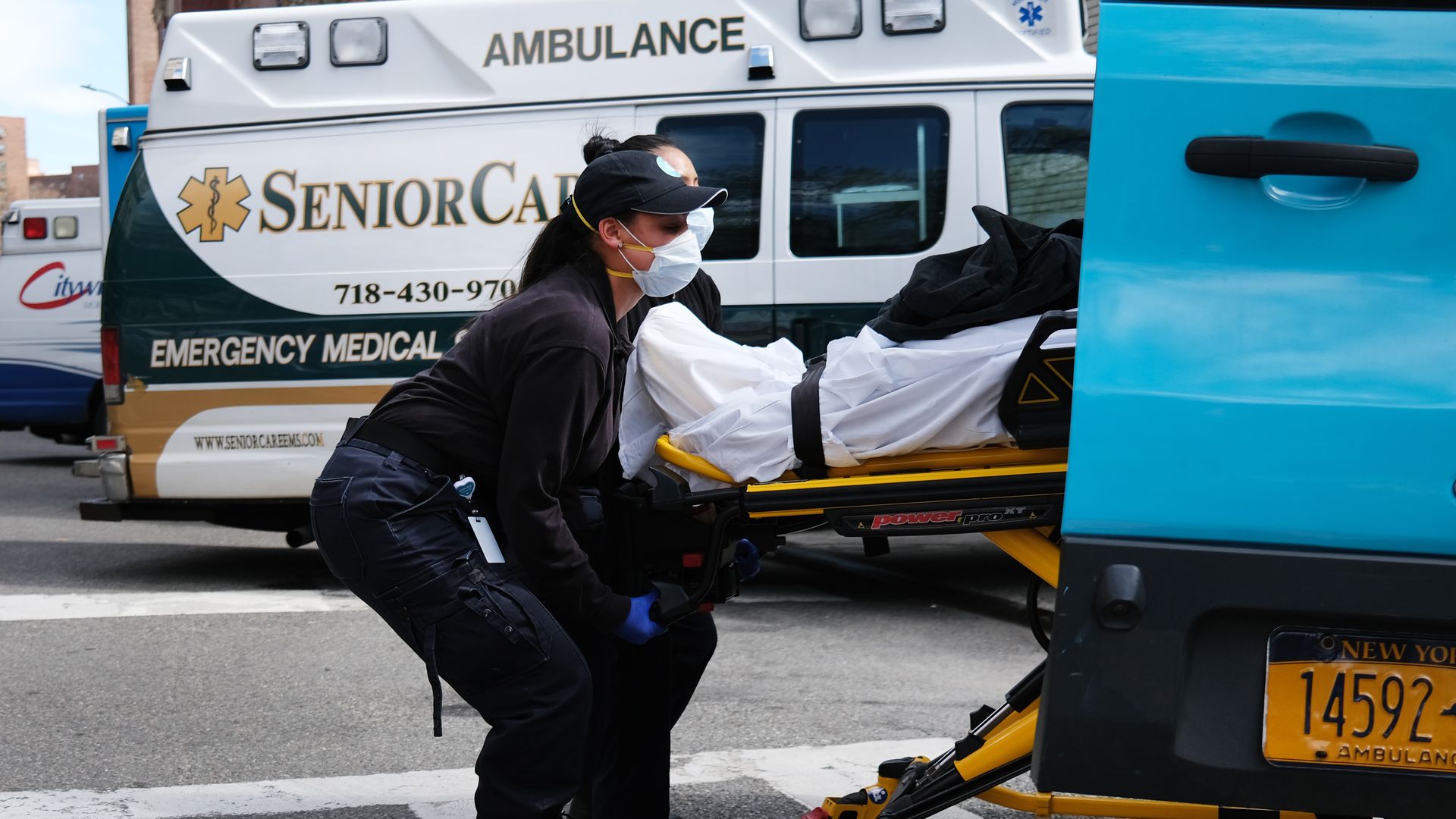 In this image, two EMTs lift a patient on a stretcher into a van