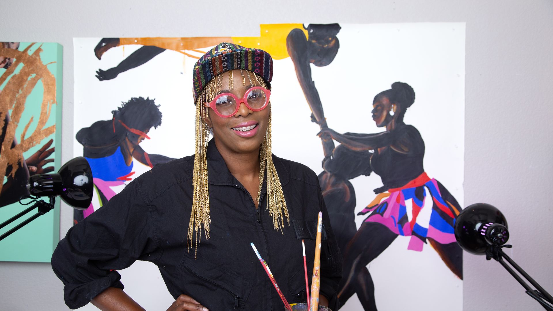Artist in a black jumpsuit, colorful cap, and pink glasses holds paintbrushes in a purple cup, standing in front of vibrant paintings of black figures dancing and moving.