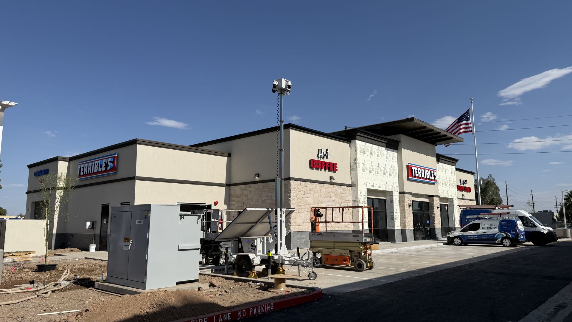 Wide view of a beige convenience store under construction against a clear blue sky; signs read "Hot Coffee" and "Terrible's"; American flag on a tall pole; construction equipment and a white van in front.