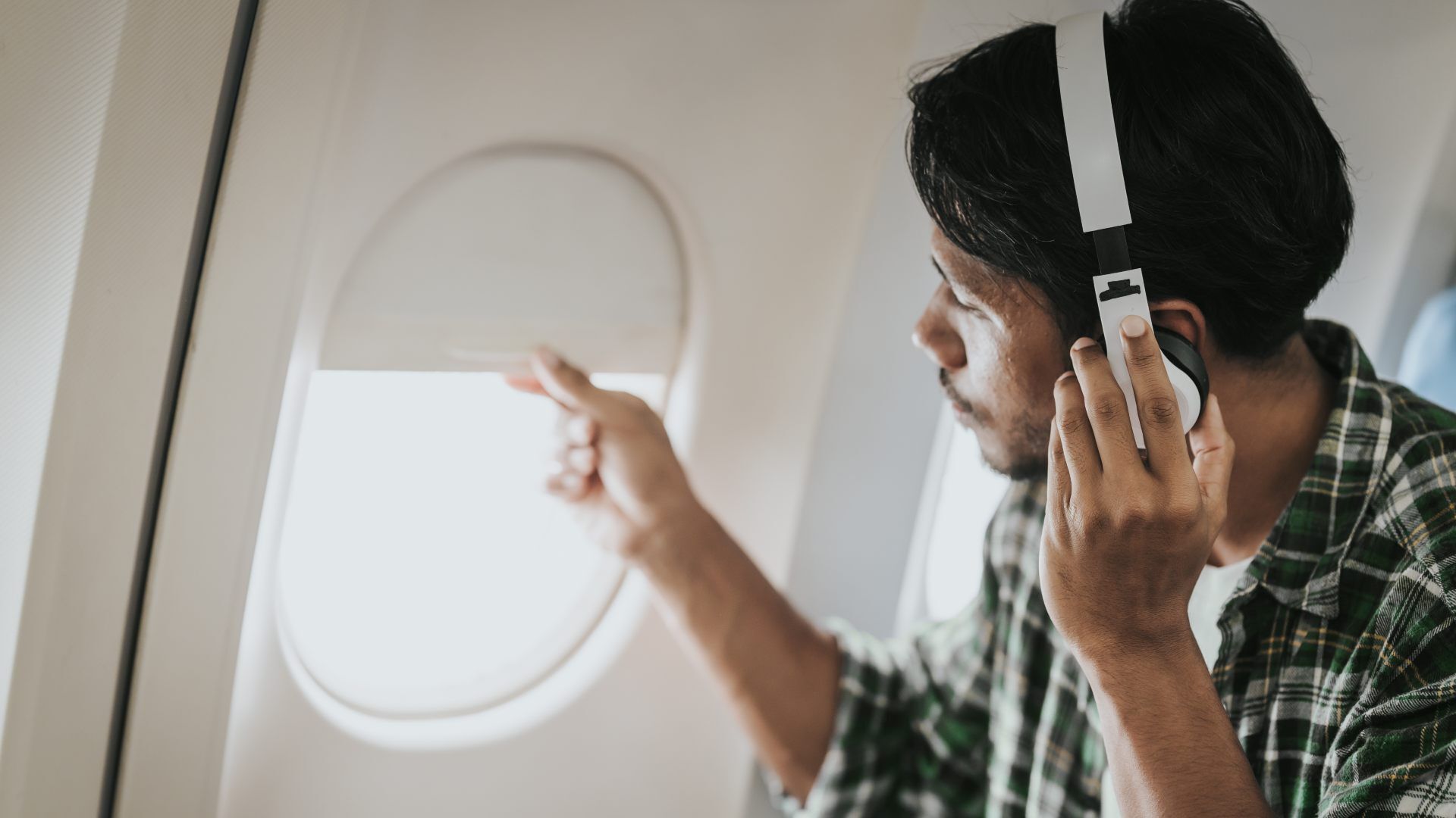 A man with dark hair wearing white over-ear headphones sits by an airplane window, looking outside and adjusting the window shade; he wears a green plaid shirt.
