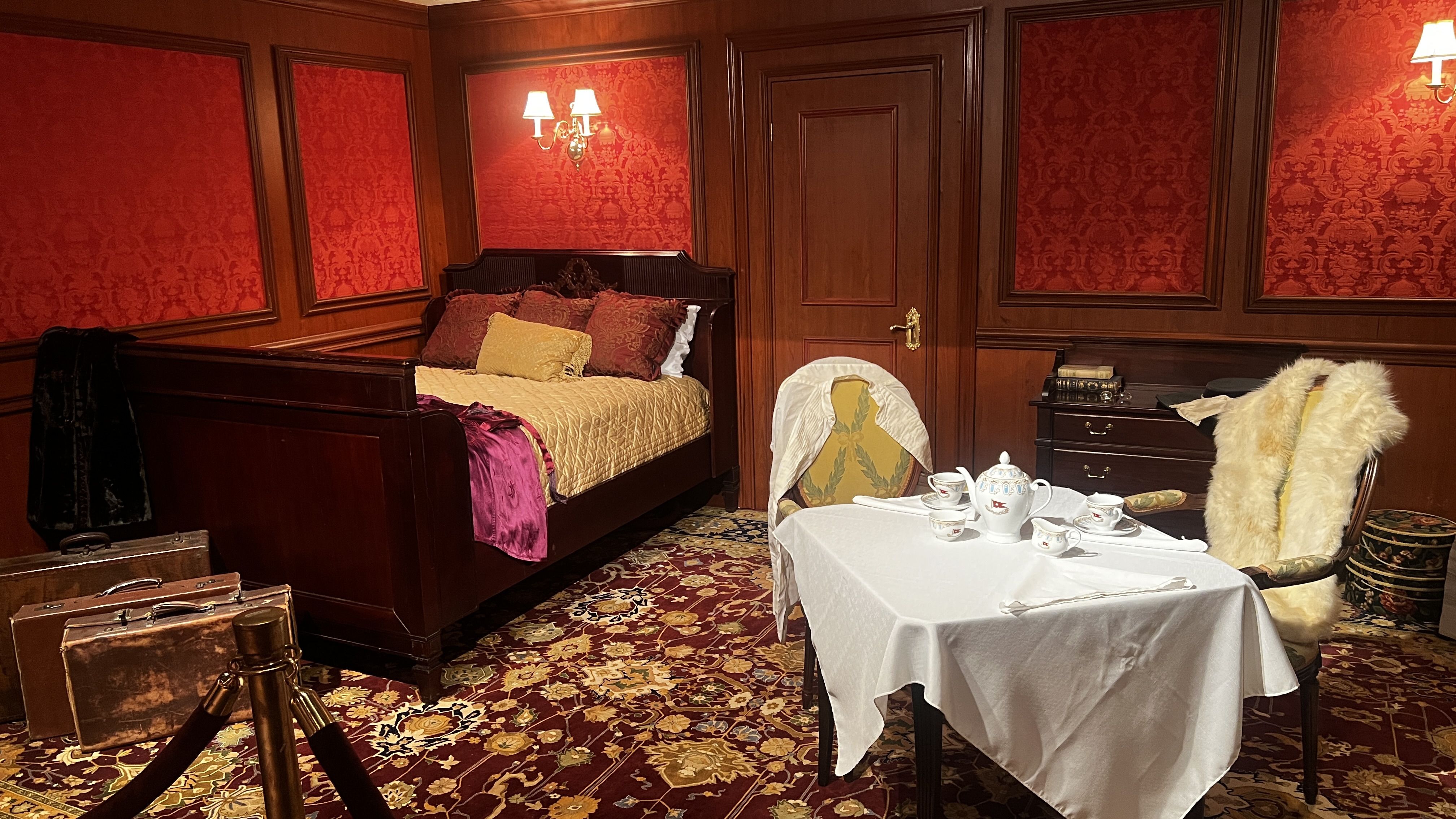 Luxurious hotel bedroom with red damask walls and dark wood paneling. A carved bed with a gold quilt and burgundy pillows, a table set with china, a fur coat on a chair, and suitcases on the floor.