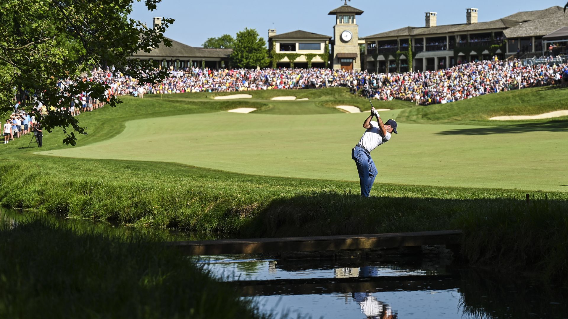 Golfer Denny McCarthy hits a golf shot near a pond and in front of a large crowd. 