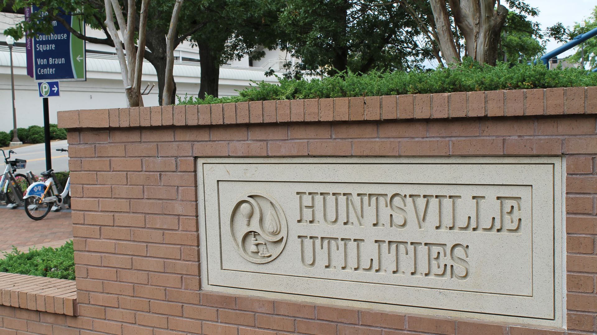 Brick wall with a stone sign reading "Huntsville Utilities" surrounded by trees and a street sign pointing to Courthouse Square and Von Braun Center in the background.