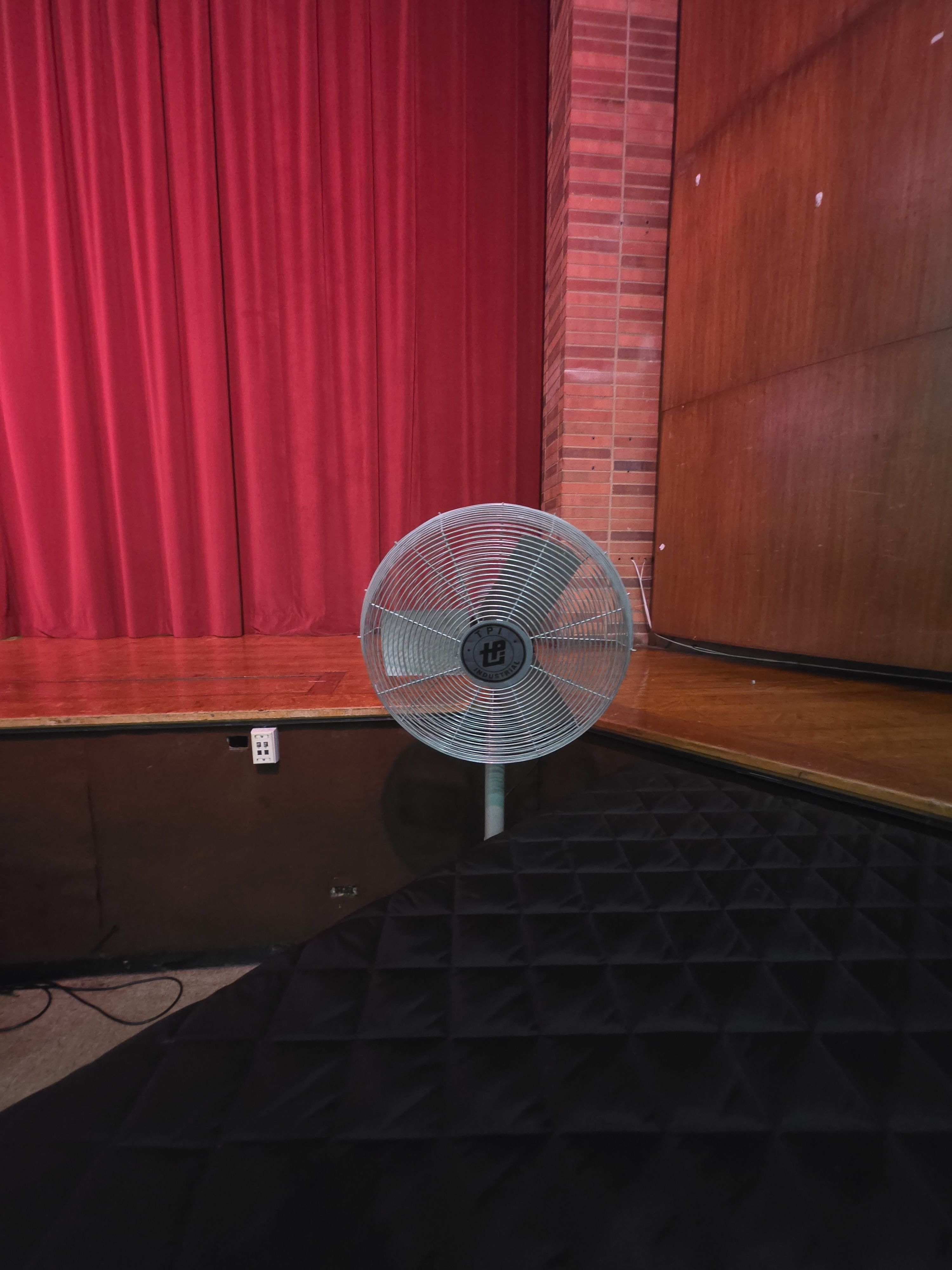 A large metal floor fan sits on the stage floor in front, with black padded steps nearby.