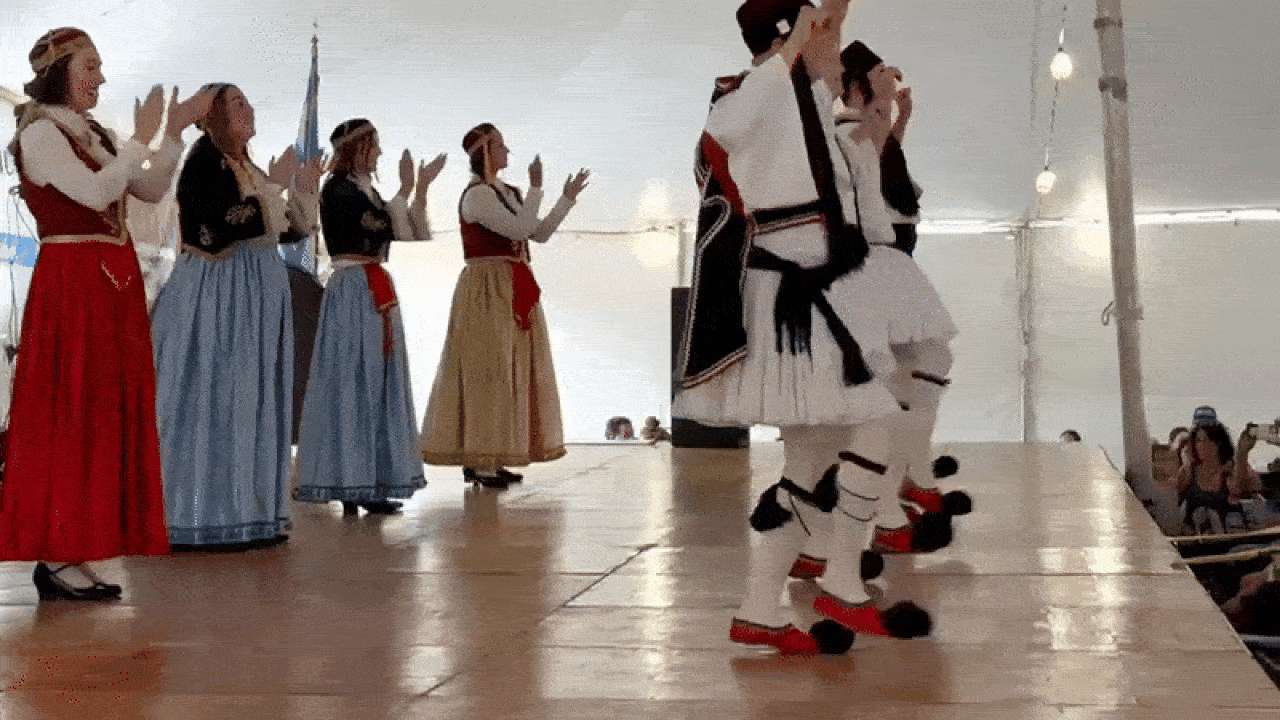 Group of men and women in traditional Greek costumes dancing on stage, with men in white skirts and black vests and women in long colorful dresses, clapping hands under a tent.