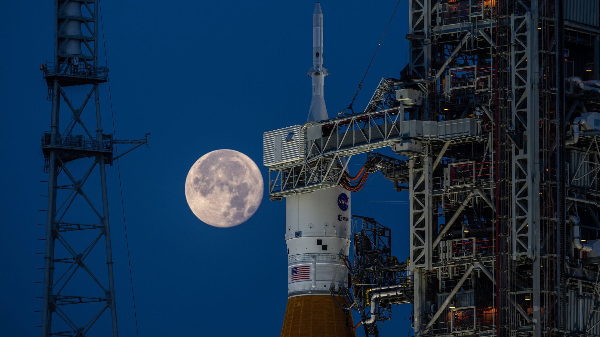 The Orion capsule atop its rocket awaiting launch with the Moon behind it in the night sky.