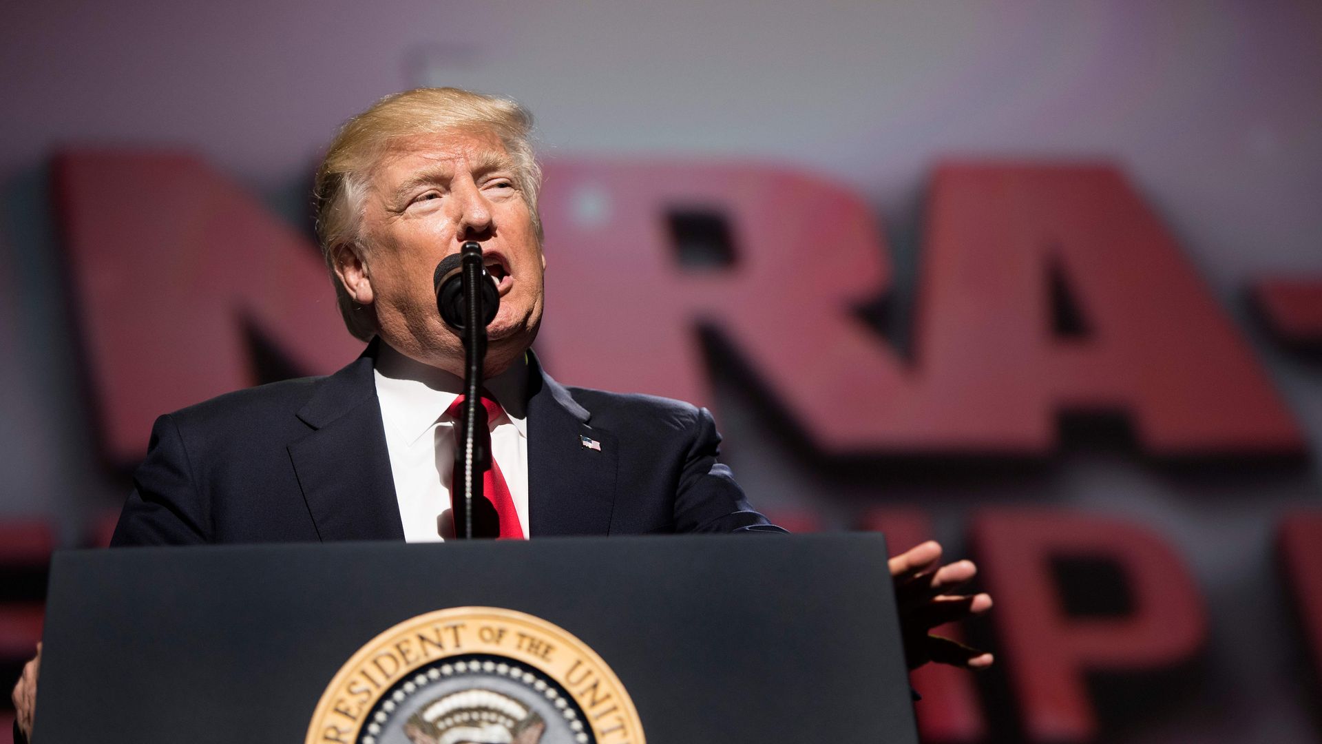 Donald Trump in a suit and red tie talking into a microphone in front of a blurred NRA sign