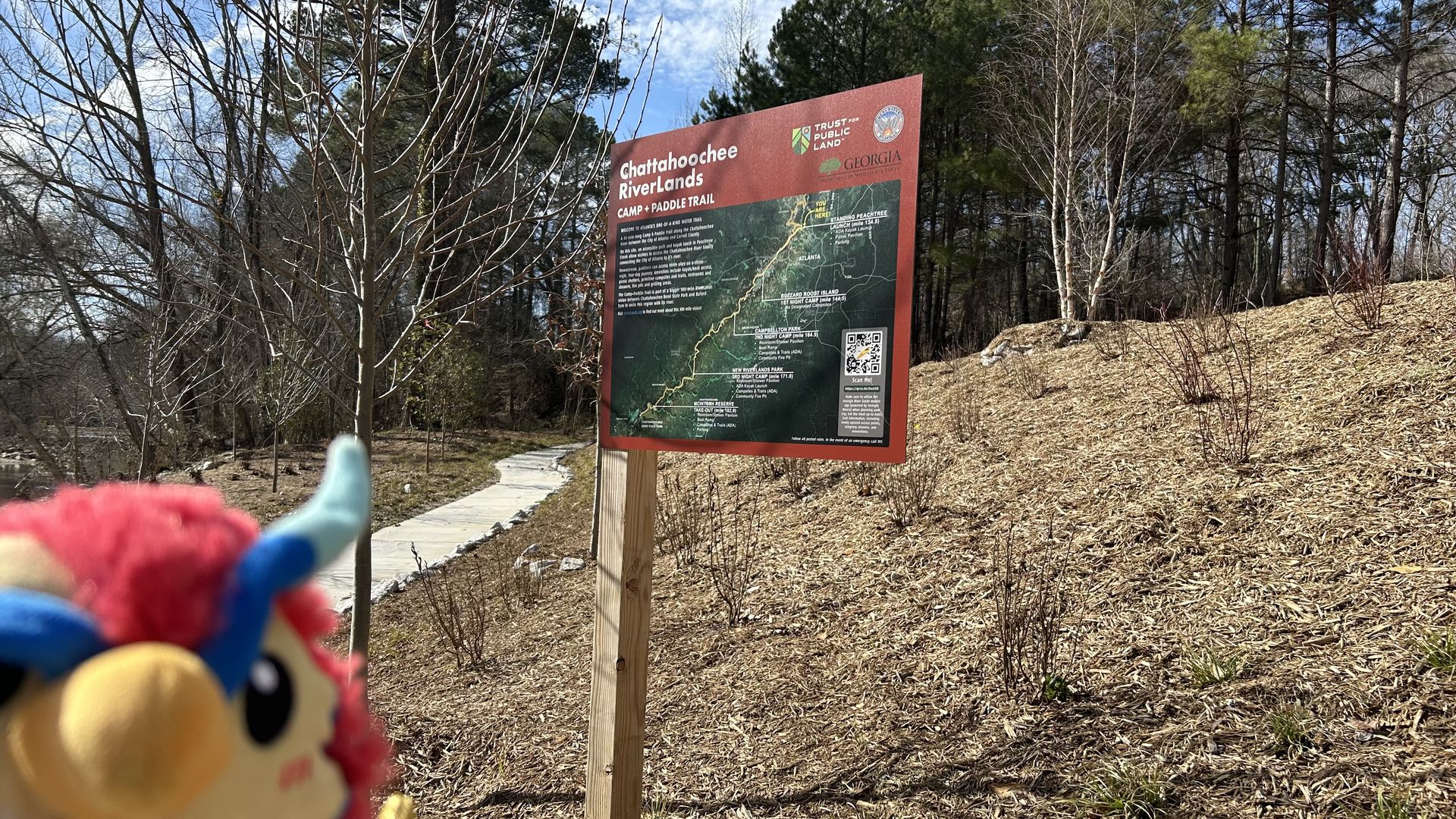 A stuffed toy animal phoenix sneaks into the bottom left of a photo of a sign with a map of a kayak trail in a wooded, sloped area
