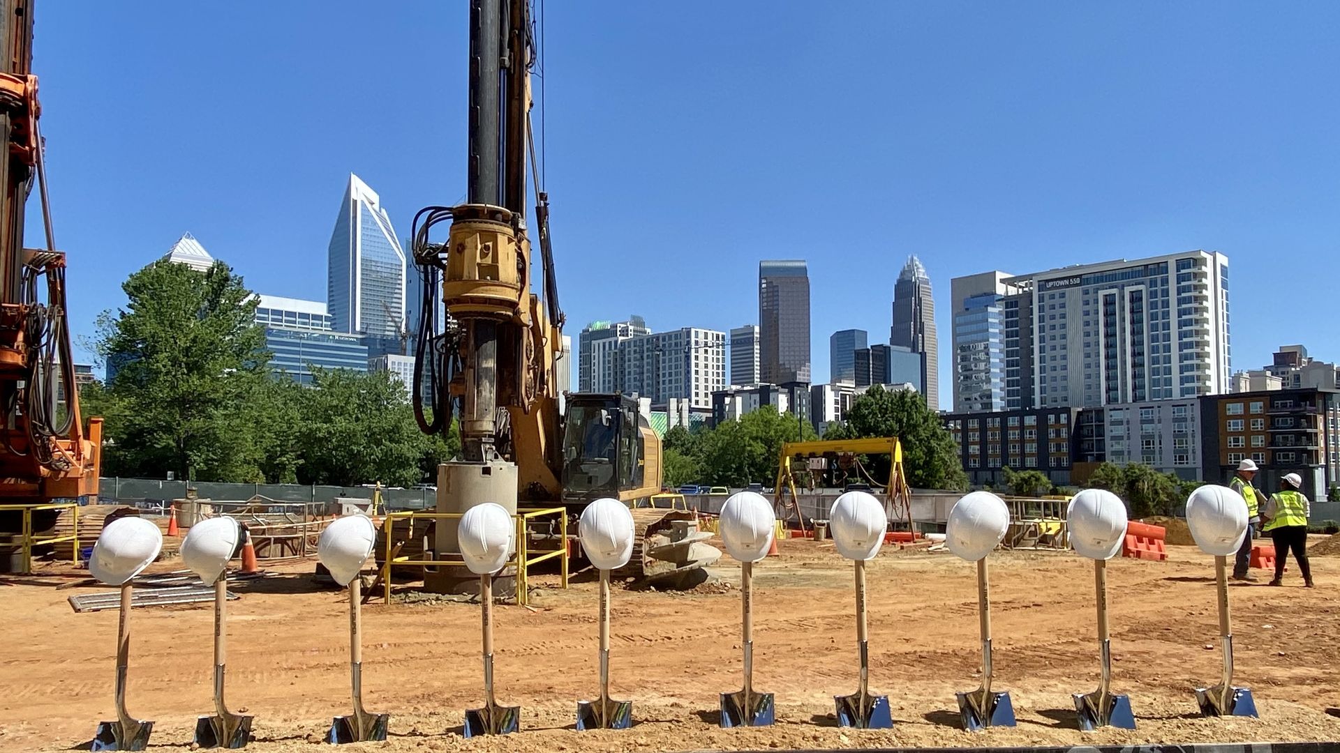 Construction site with 10 shovels lined up stuck in the sand and 10 hard hats stacked on top. There's a crane in the background and behind that is the Charlotte skyline. 