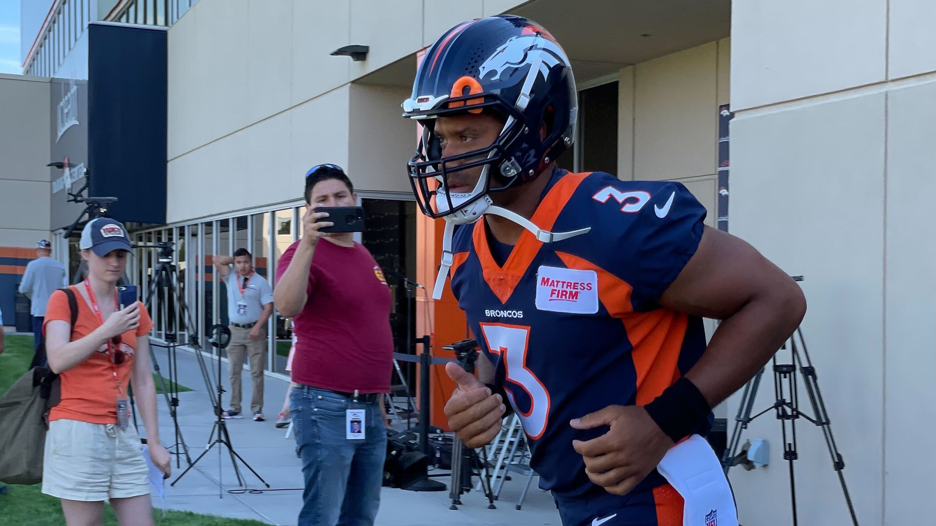 A man in an navy blue American football uniform with orange lines runs past people taking photographs.m 