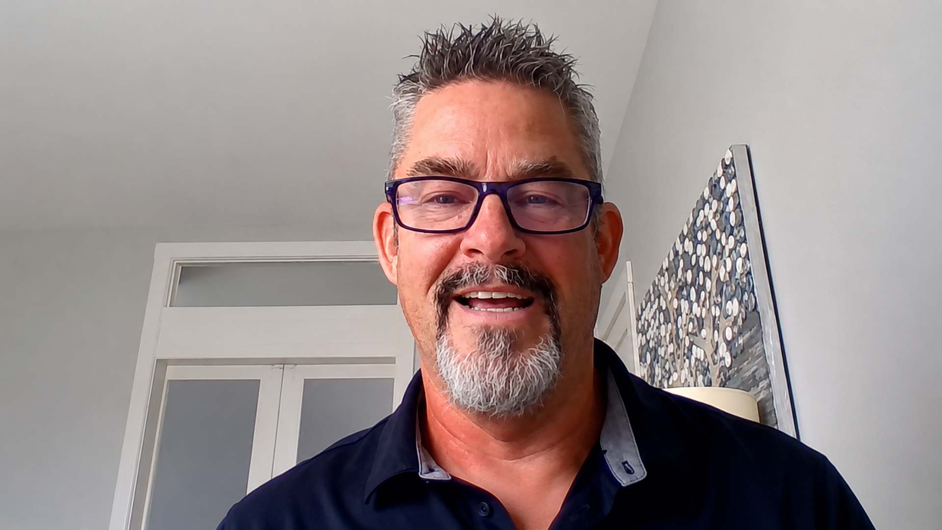 A man with spiky salt-and-pepper hair, purple glasses, and a gray goatee smiles at the camera. He wears a dark polo shirt, with a circular-pattern art piece on the wall behind him.