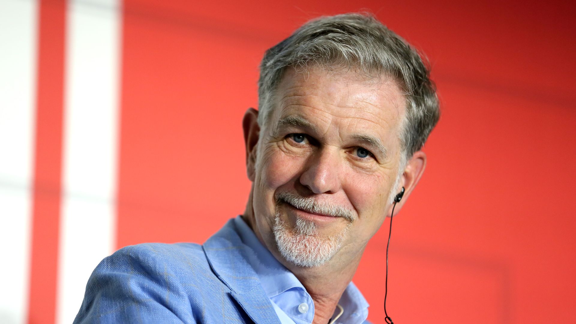 Reed Hastings in a light blue suit and a light blue shirt sits in front of a red backdrop.