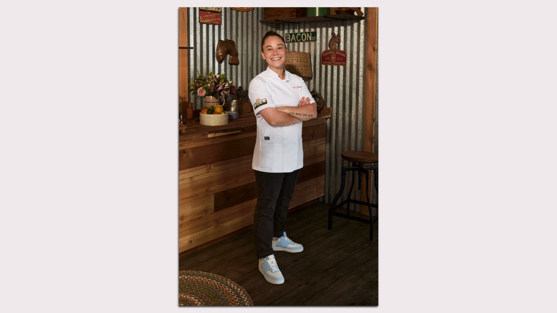 Person in a white chef coat, black pants, blue and white sneakers, standing with arms crossed in a rustic kitchen with wooden and corrugated metal walls, smiling.
