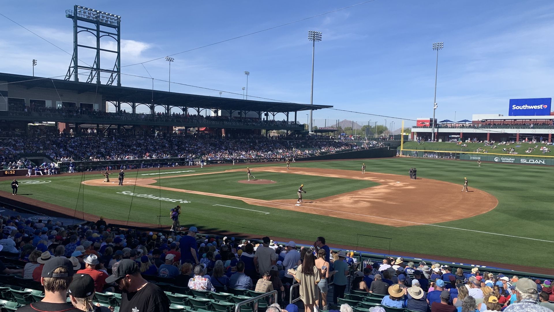Sunny baseball stadium with a crowd watching players on field. Green grass, brown infield, and blue sky. "Southwest" sign on right scoreboard and infield text.