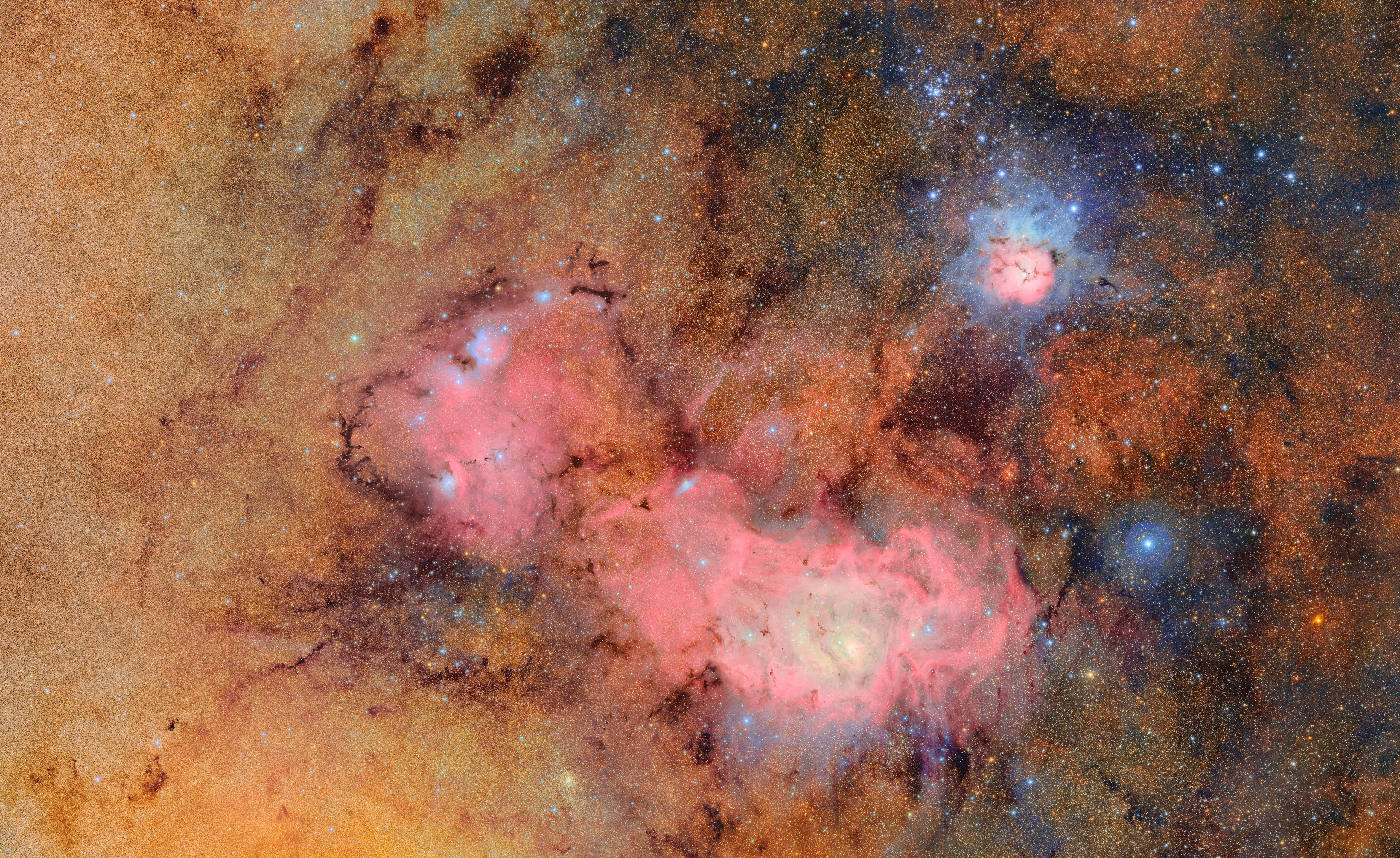 A vibrant image of a star-filled region of space featuring the Lagoon Nebula and Trifid Nebula. The scene is rich with colorful interstellar dust clouds and bright stars set against a dark backdrop.