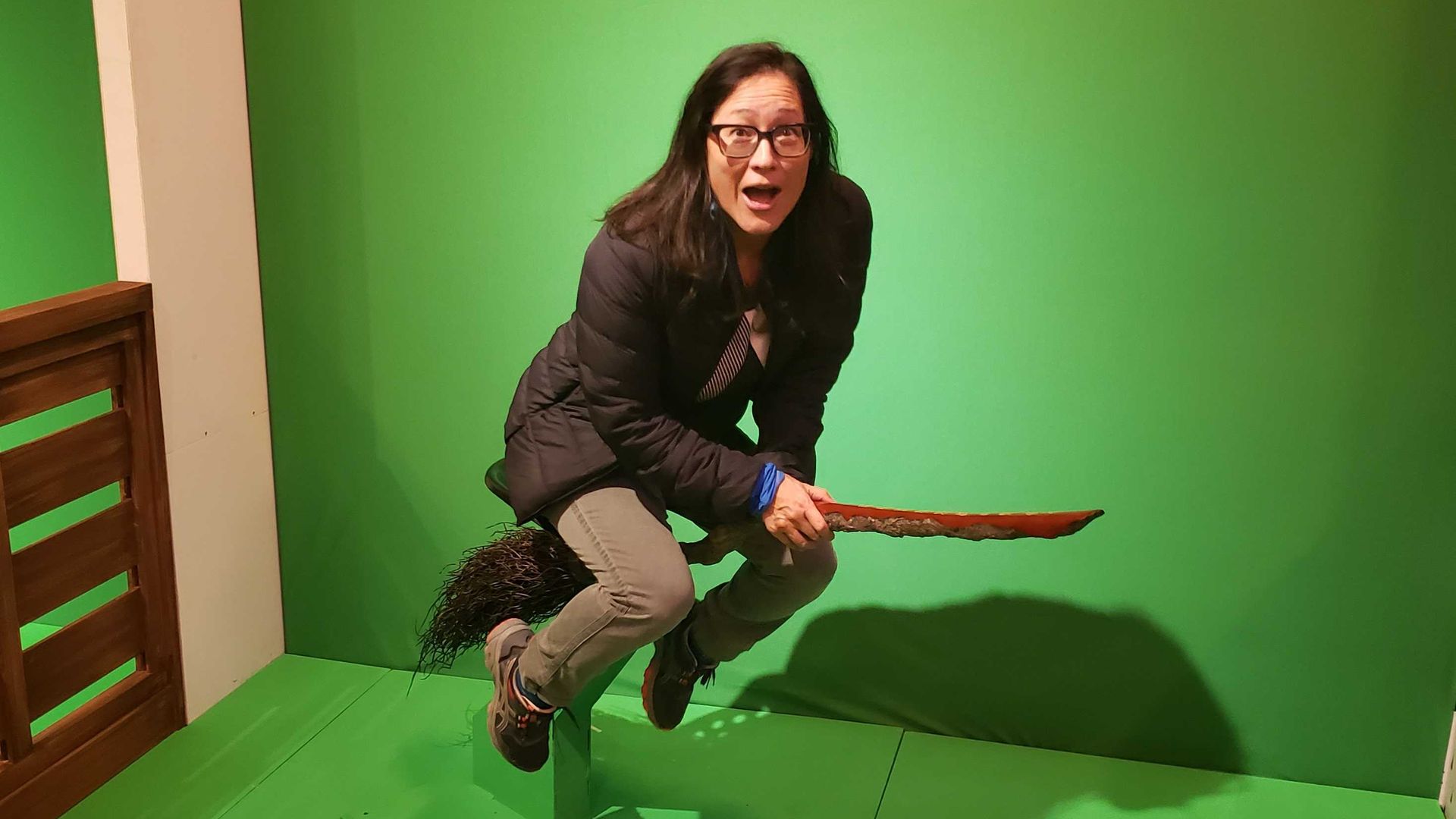 Photo of a woman on a broom with a green screen behind her. 