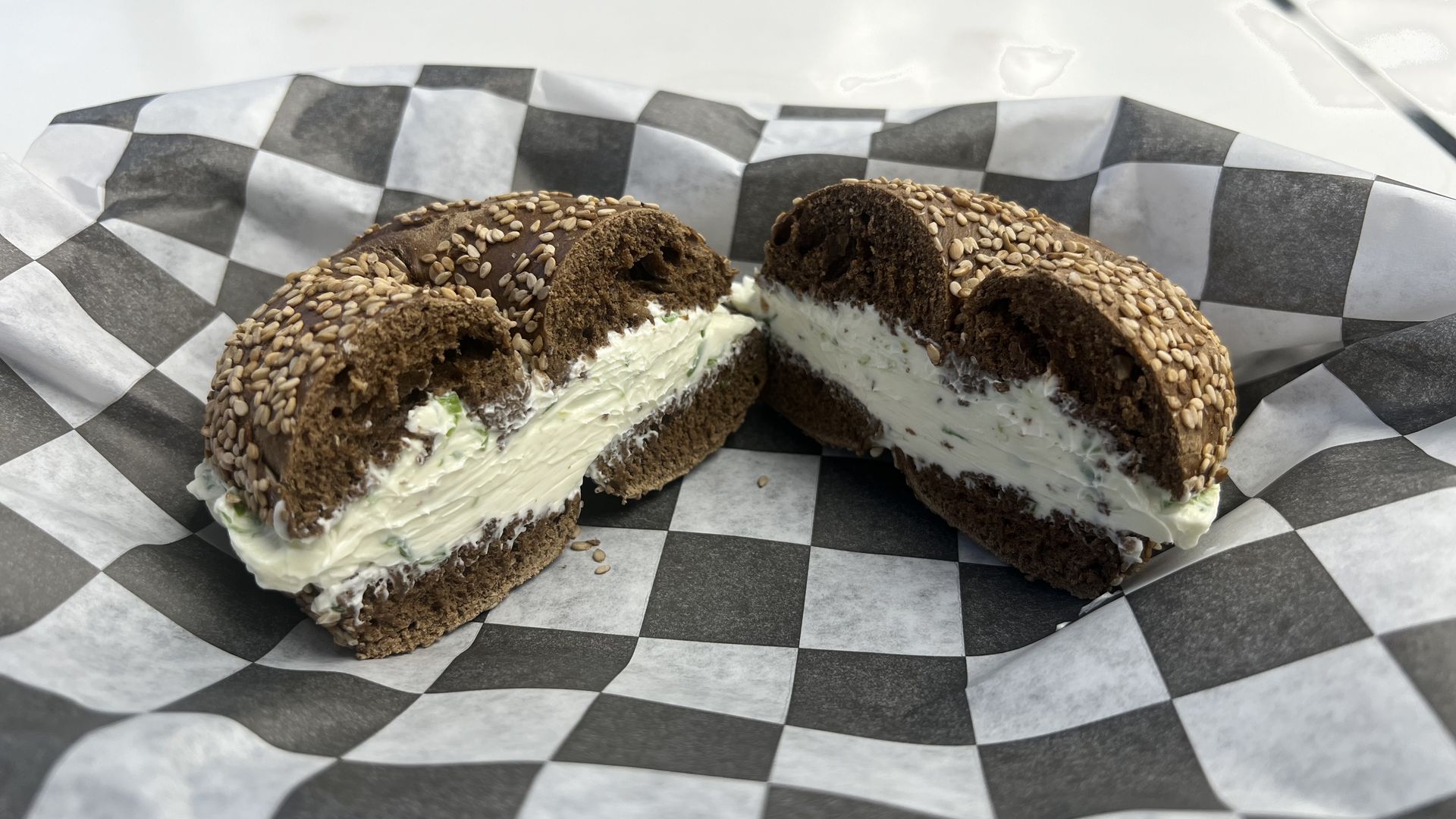 Two sesame-seed bagel halves filled with creamy herb cheese on a black-and-white checkered paper liner.