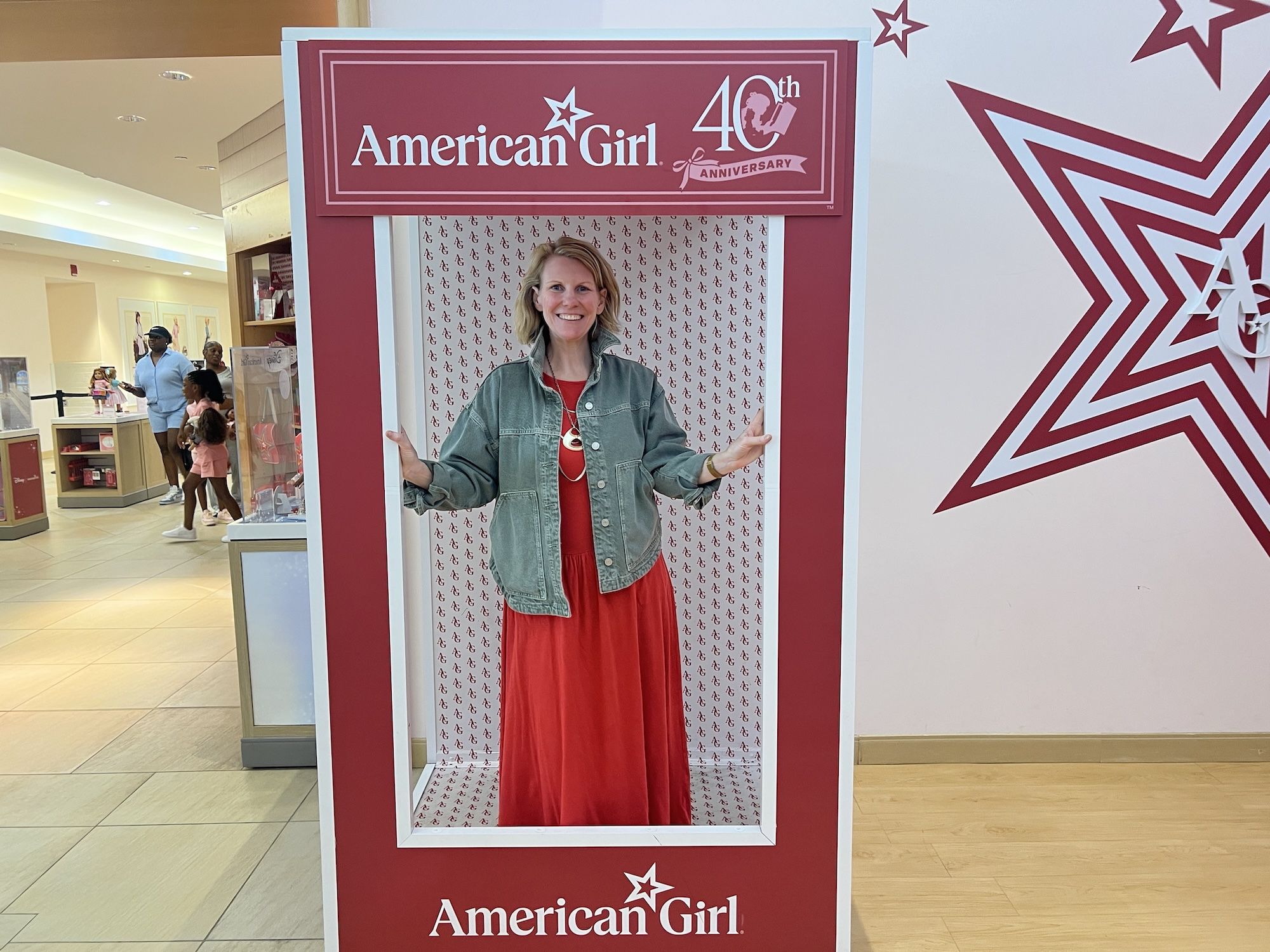 Smiling woman in a red dress and denim jacket stands inside a maroon "American Girl" photo frame with a "40th anniversary" emblem, in a mall display with children nearby.