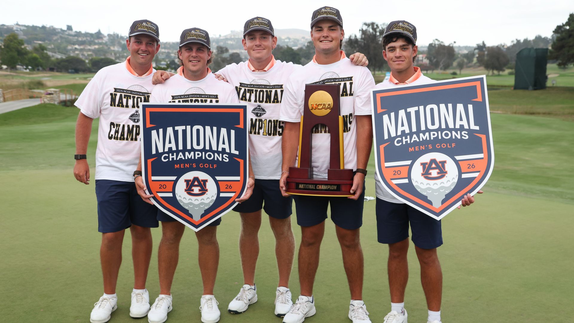 Five golfers in white shirts and caps pose on a golf course, smiling, holding large National Champions signs for men's golf and a tall trophy between them, with a scenic hillside background.