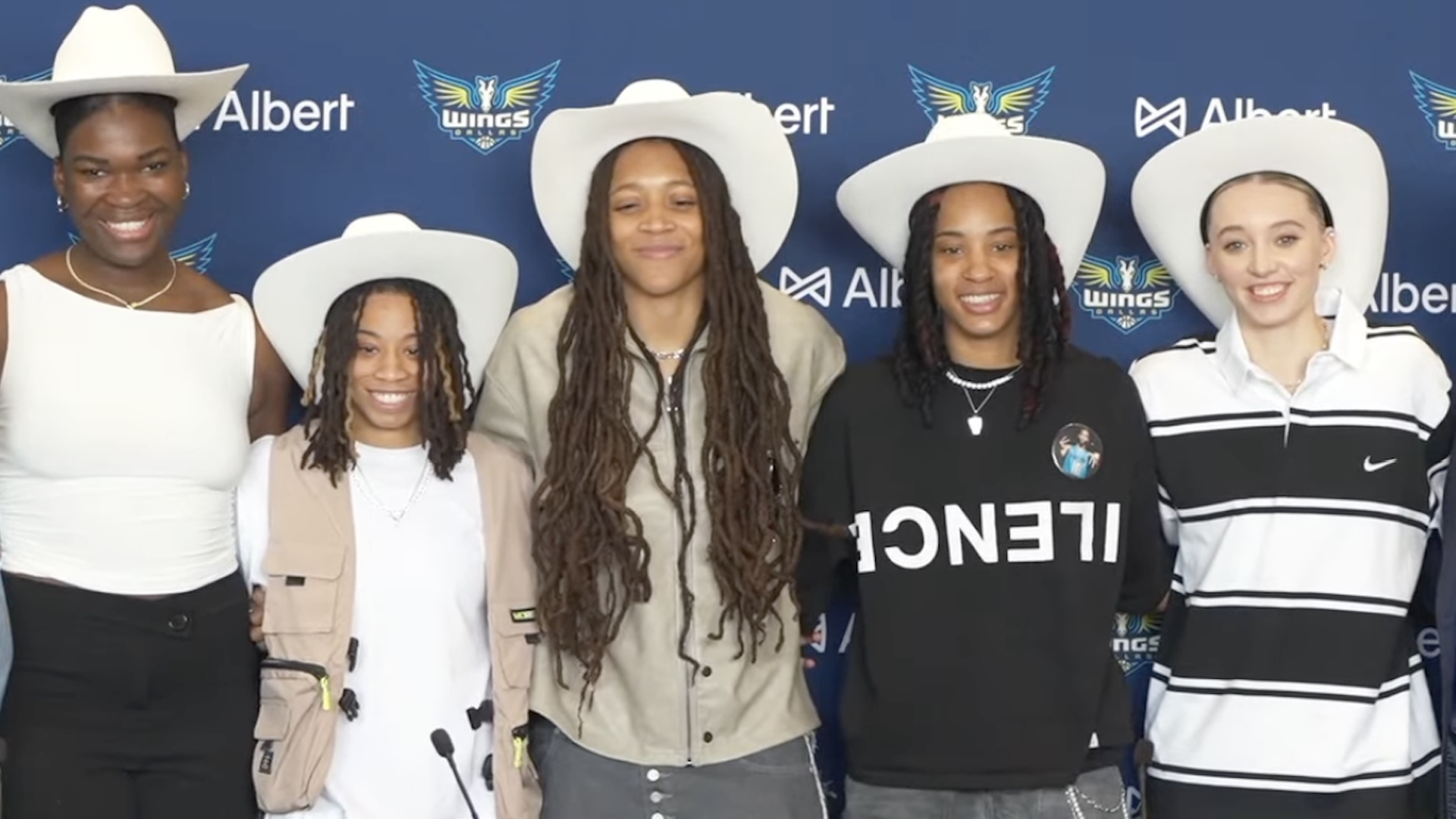 Five women stand in a line, wearing white cowboy hats