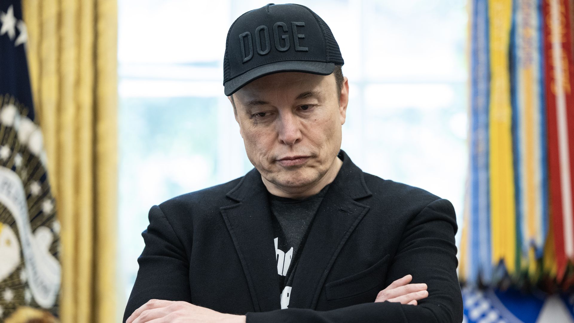 Elon Musk wearing black blazer and a black "DOGE" hat, standing in the Oval Office with his arms crossed.