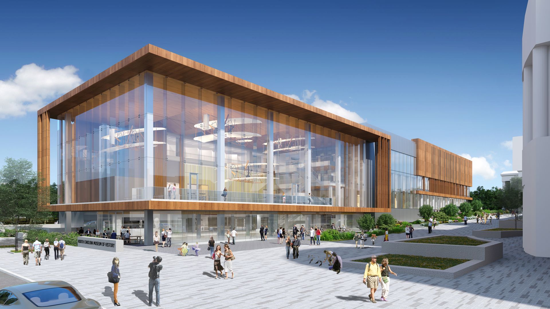 Modern building with large glass walls and wooden accents labeled North Carolina Museum of History, people walking and interacting outside under a clear blue sky.