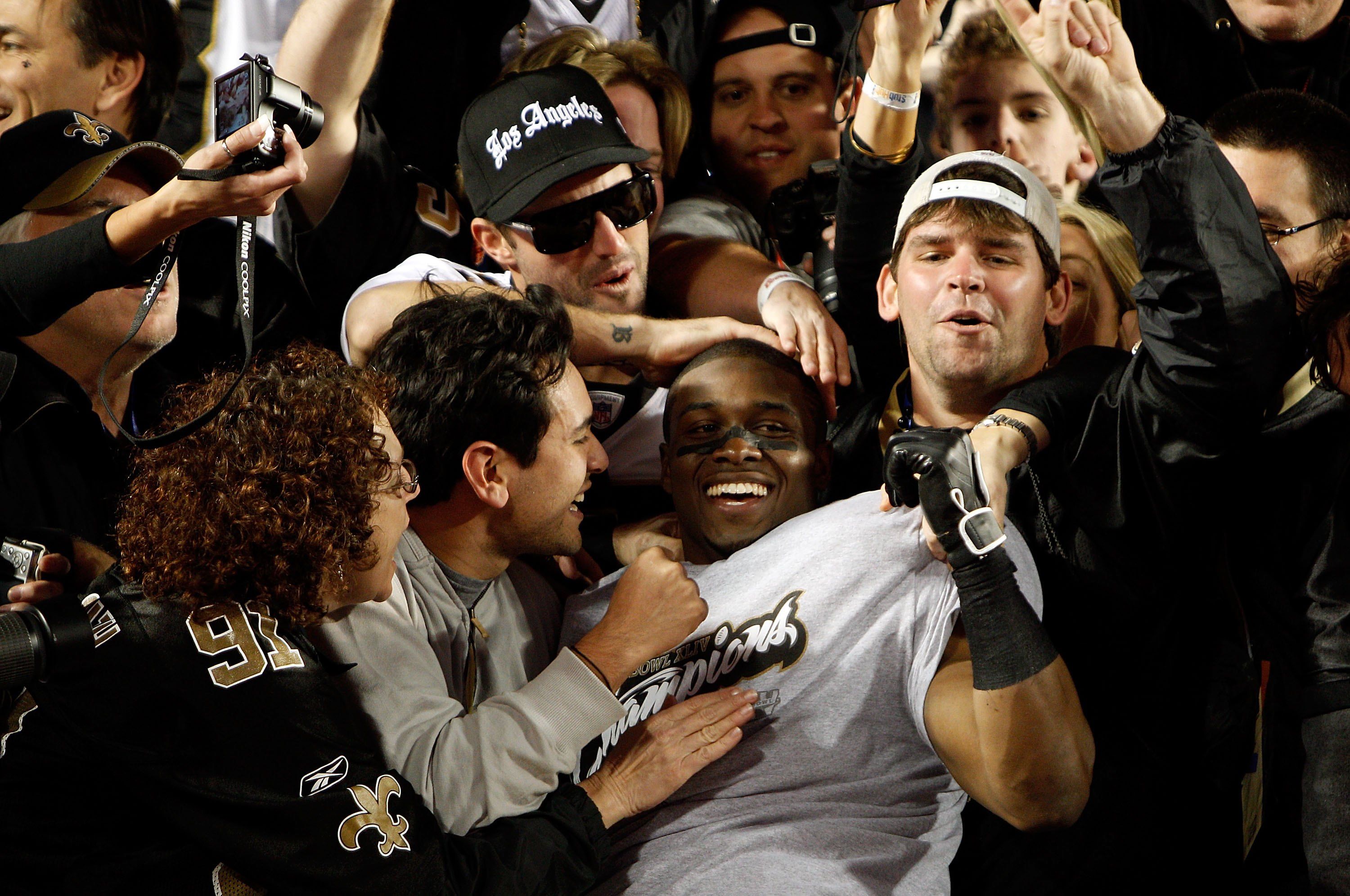 Smiling football player wearing a gray "Champions" shirt celebrates with cheering fans, some in black and gold New Orleans Saints gear, capturing a joyful victory moment on the field.