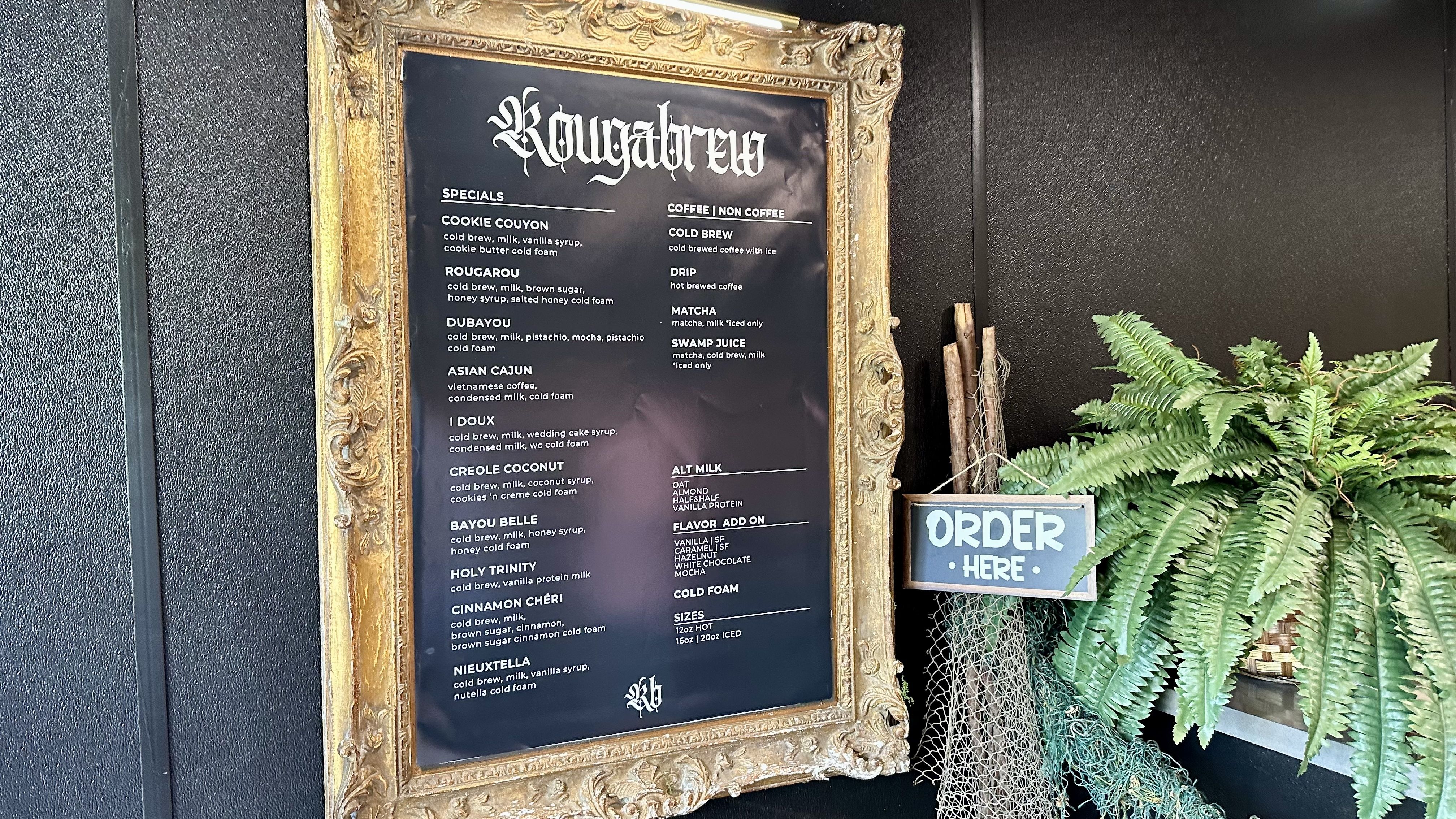 An ornate gold frame houses a black menu board with white gothic text, mounted on a dark textured wall; a blue 'ORDER HERE' sign and lush green ferns sit to the right.
