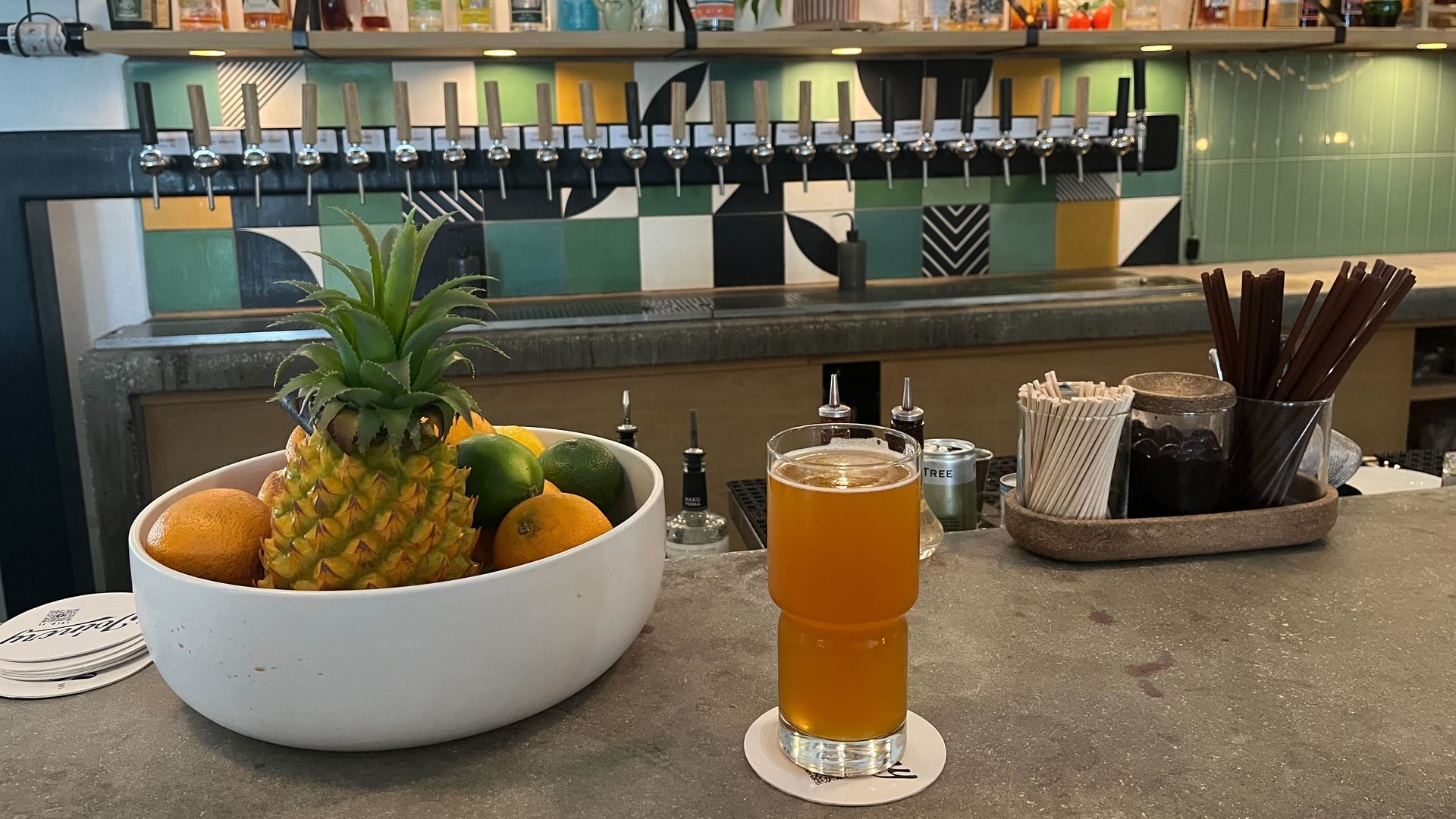 A pint of orange-colored beer next to a bowl of fruit. Beer taps jut out from a wall decorated with green, yellow, black and white tiles.