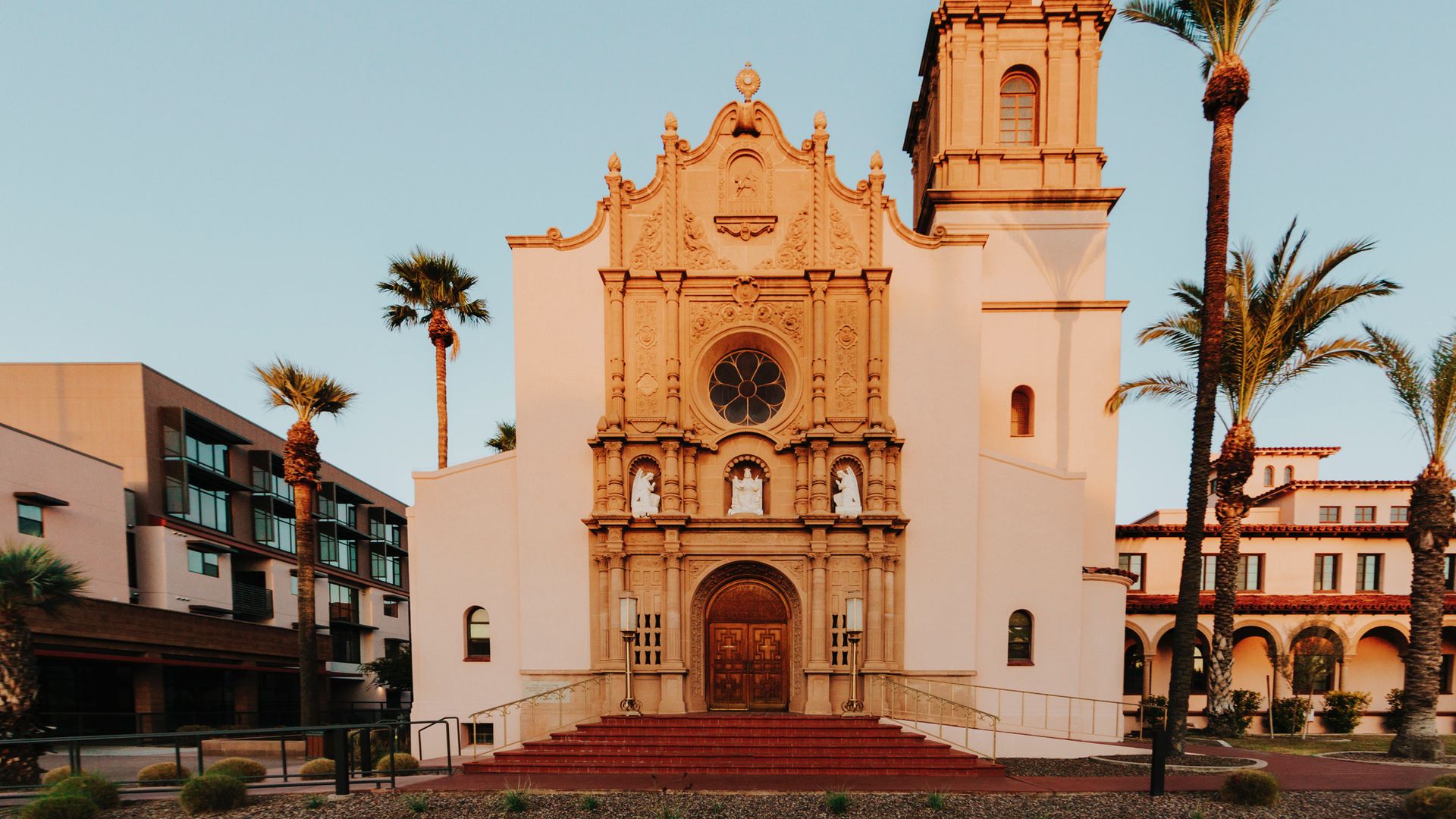 Sunlit historic church with ornate tan facade, large wooden doors, and a bell tower topped with a cross, surrounded by palm trees and modern buildings under a clear sky.