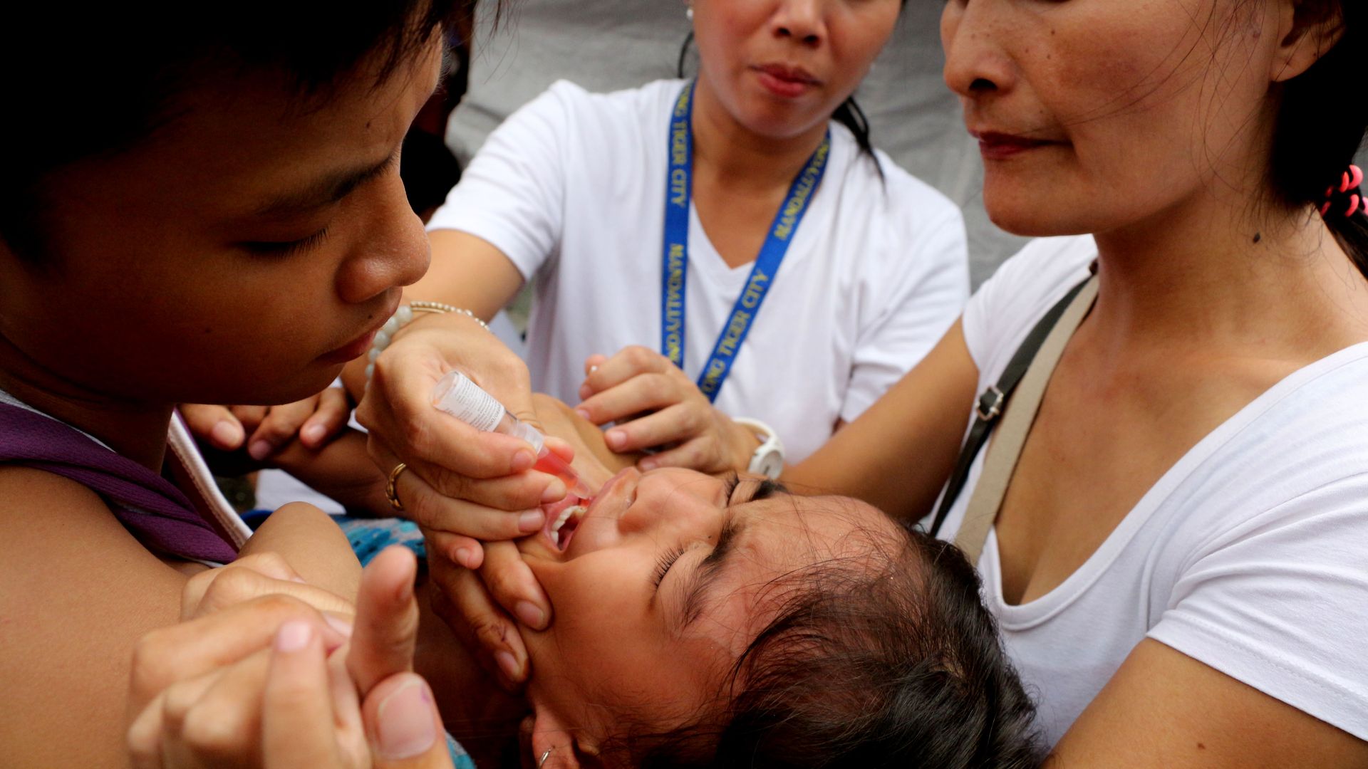 In this image, three women hold a child as a vaccine is administered to it through a dropper into its mouth.