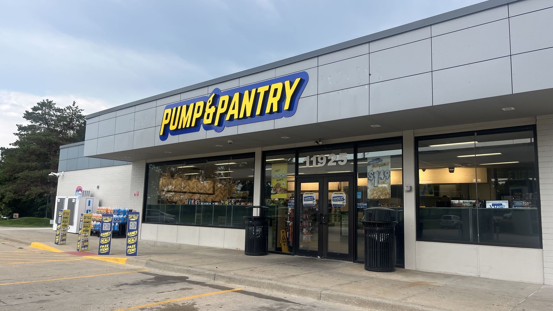 Exterior view of a Pump & Pantry convenience store with a large yellow and blue sign, gray panel walls, glass entry doors, promotional boards outside, and trees to the left under a cloudy sky.
