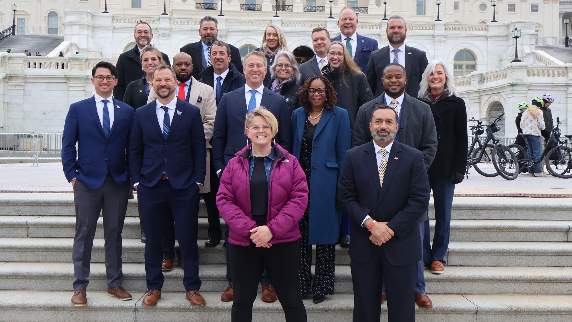 Group of diverse professionals in suits posing on the steps of a white neoclassical government building; cyclists stand to the right, and a woman in a magenta jacket is center-front.