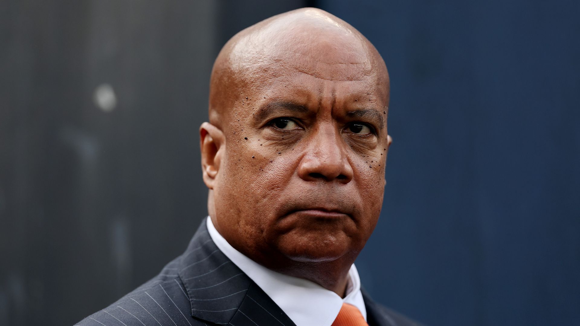 Close-up of a serious bald man wearing a dark pinstripe suit, white shirt, and bright orange tie against a blurred dark blue and gray background.