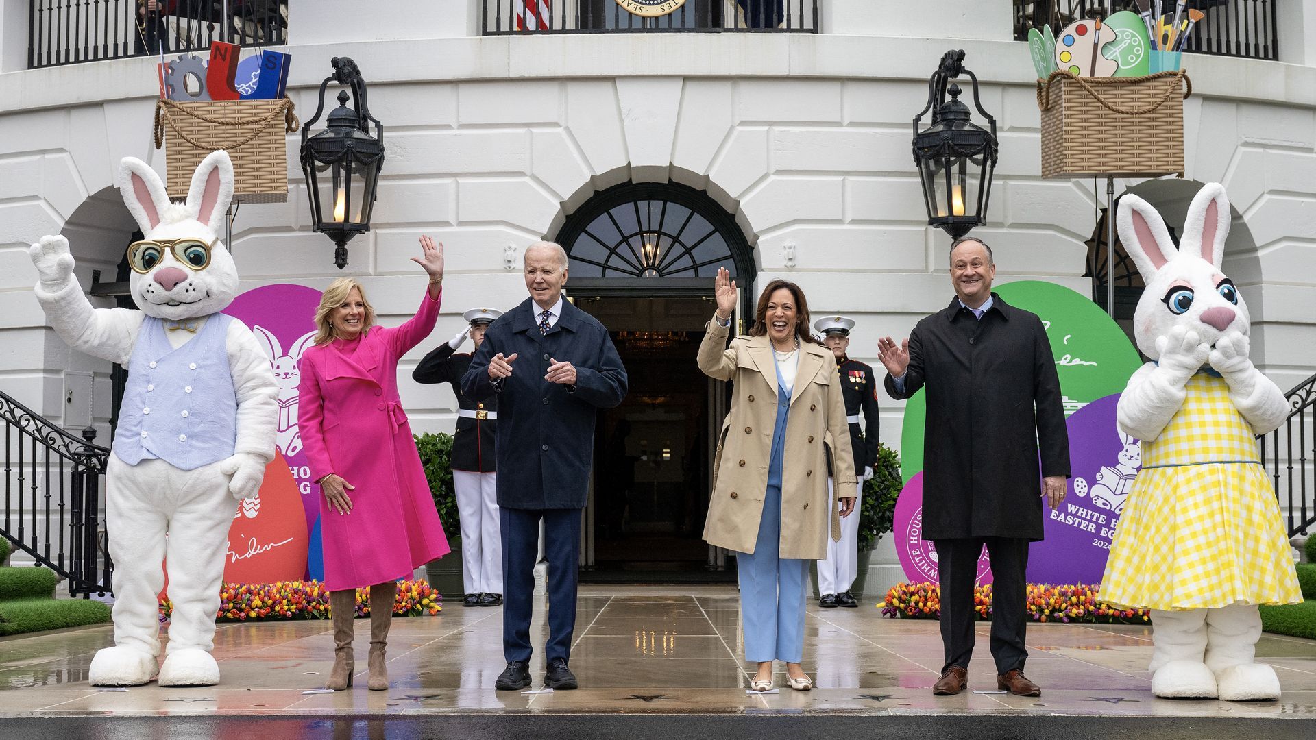 From left to right, Jill Biden, wearing pink, Joe Biden, wearing a dark suit, Kamala Harris, wearing a blue jumpsuit and Doug Emhoff, wearing a dark suit, stand and wave to an audience. To either side is a large plush Easter bunny. 