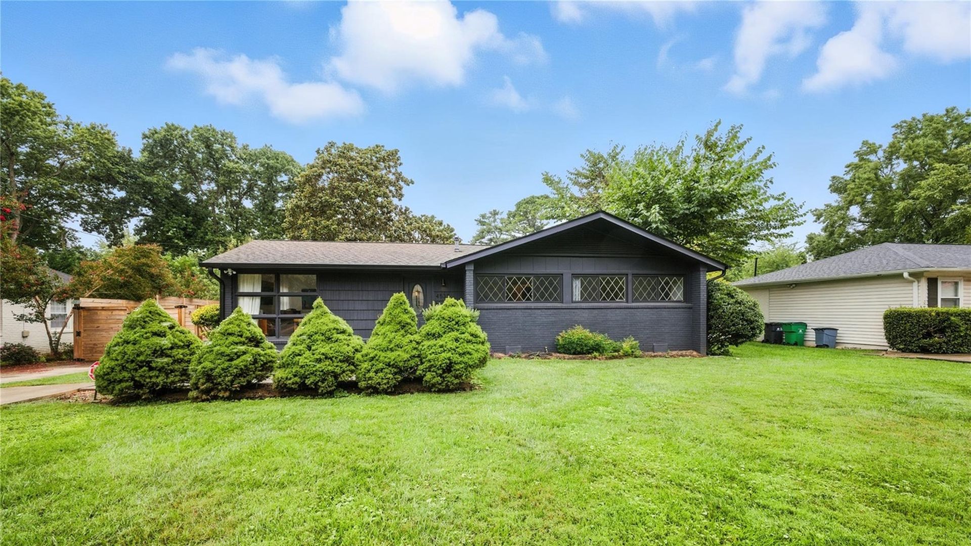 Single-story dark gray house with a triangular roof, diamond-patterned windows, a green lawn, and five trimmed conical bushes in front under a partly cloudy blue sky.
