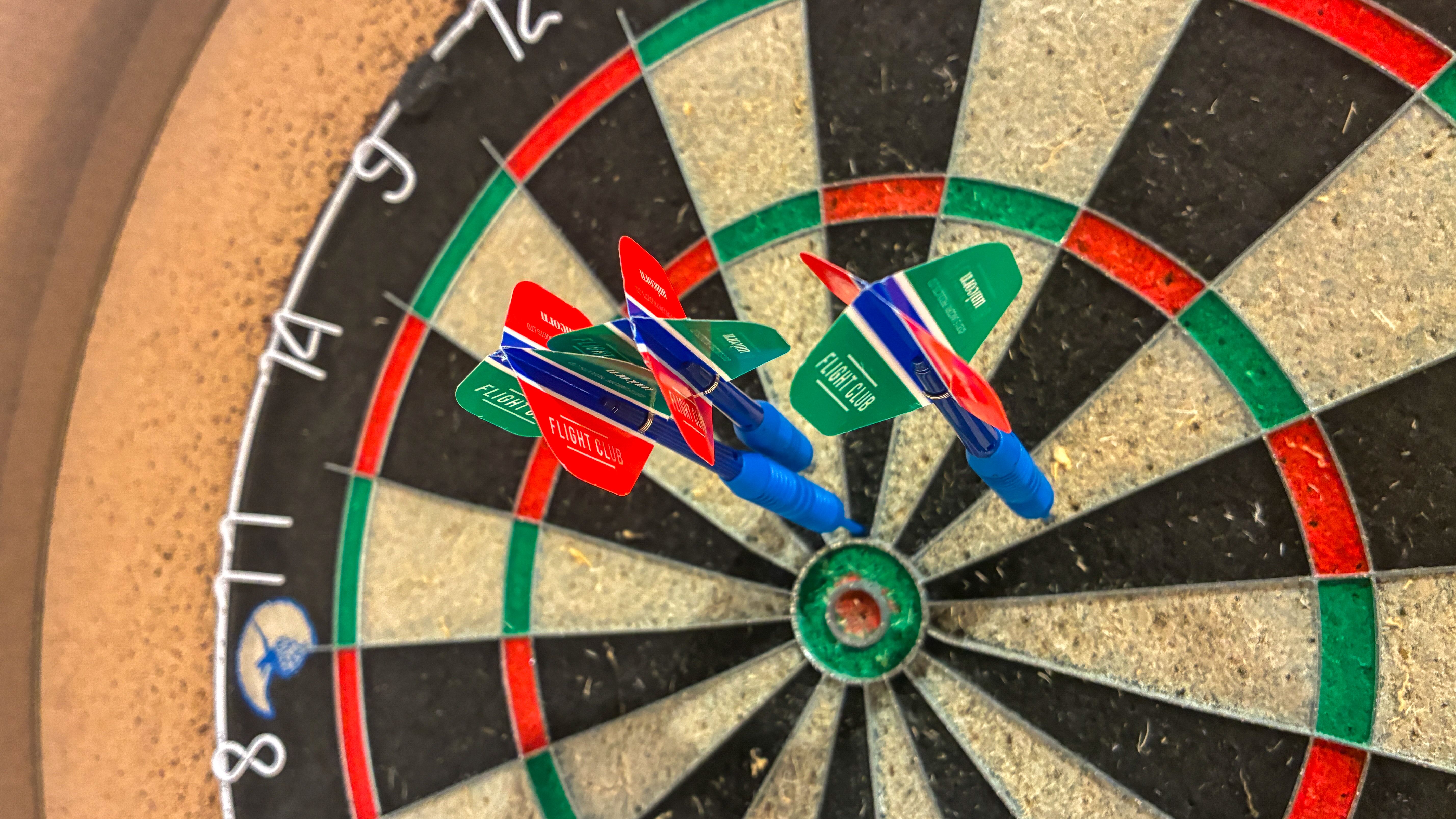Close-up of a dartboard showing a cluster of blue darts with red and green flights near the bullseye, surrounded by multicolored red, green, and black segments.