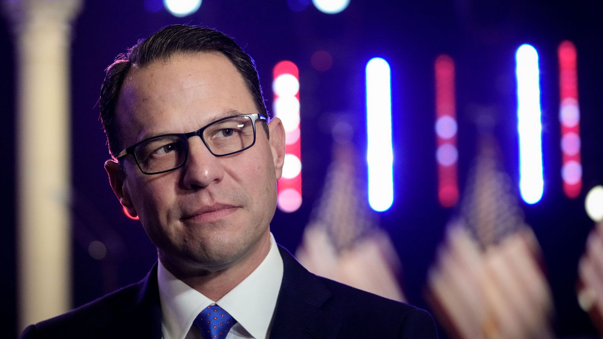Josh Shapiro, wearing a dark blue suit and standing in front of blue and red lights and American flags.