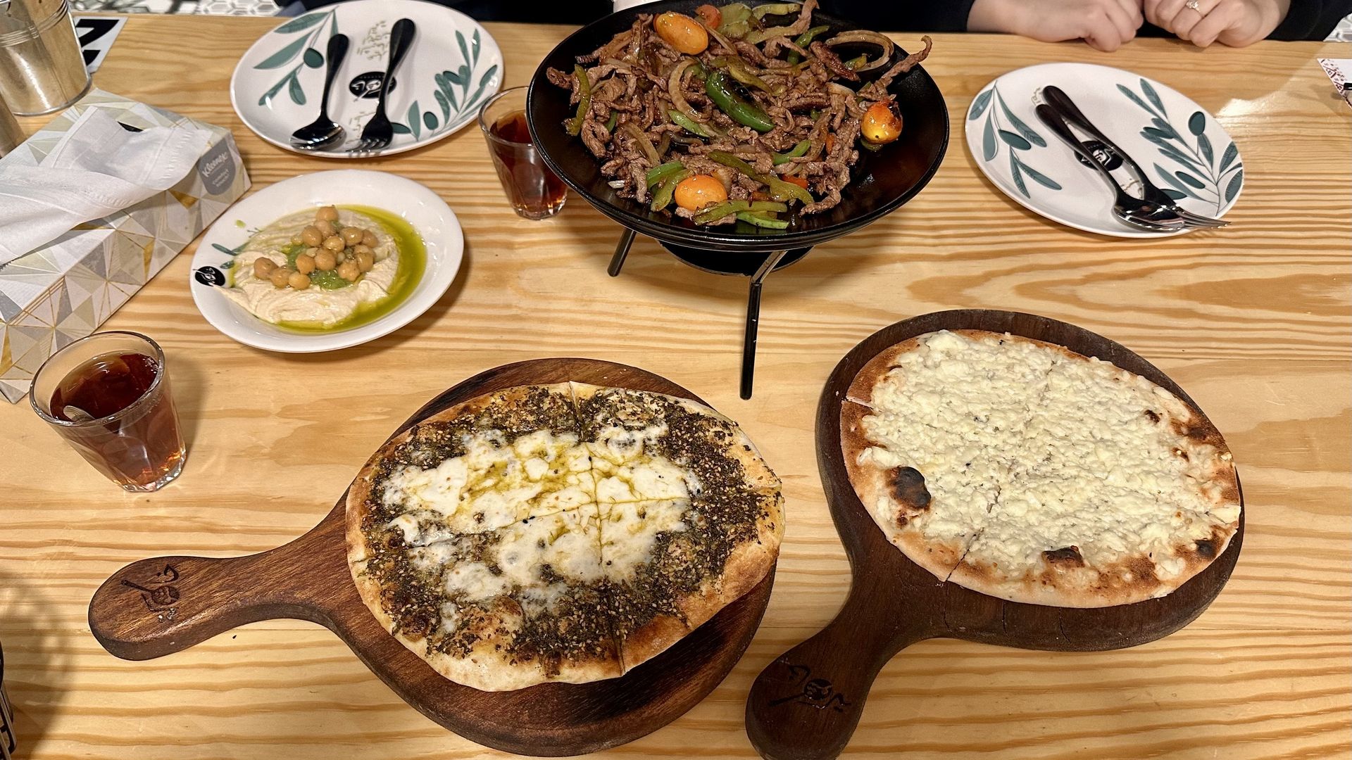 Table with two flatbreads, one topped with za'atar and cheese, the other with white cheese, a dish of hummus and a bowl of beef stir-fry with peppers