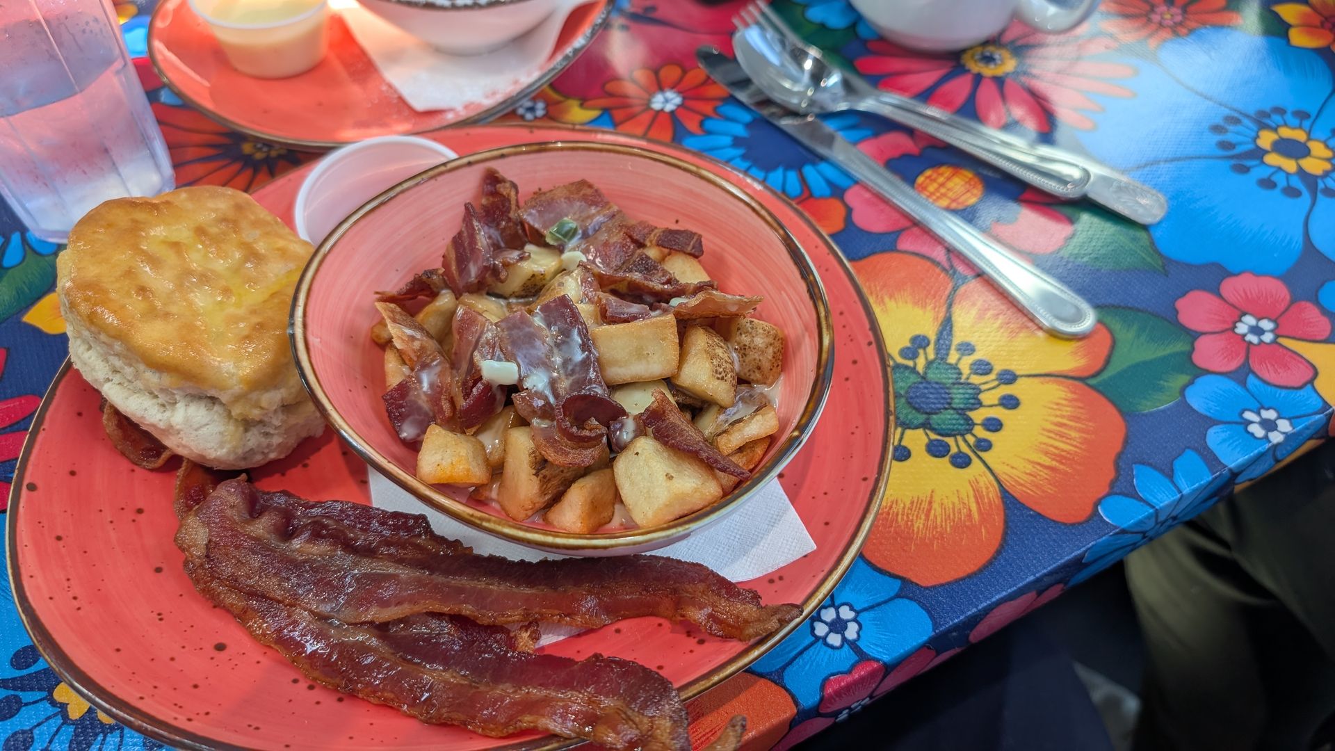 Pink plate with crispy bacon, a bowl of hash browns with bacon bits, a golden biscuit, on a bright floral tablecloth; silverware nearby.